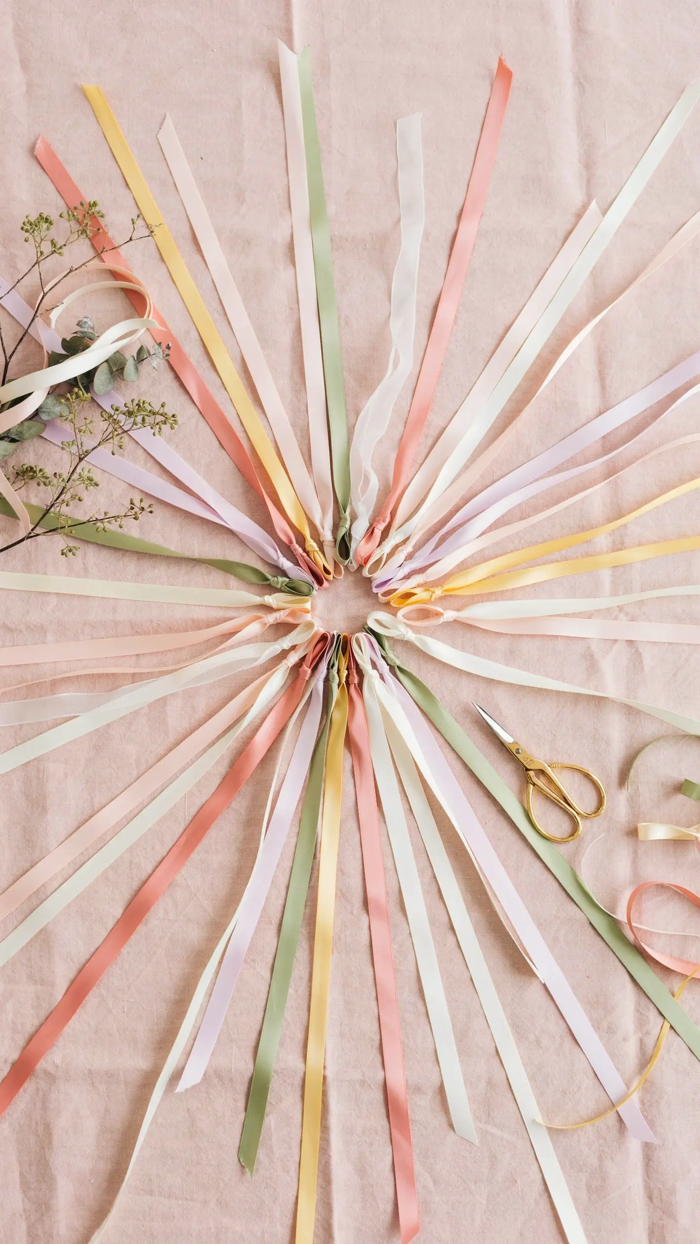 A flat lay of pastel peach, cream, yellow, and sage green ribbons fanned out in a starburst shape with gold scissors and eucalyptus on a blush fabric for a "Measure Mommy's Belly" baby shower game. 