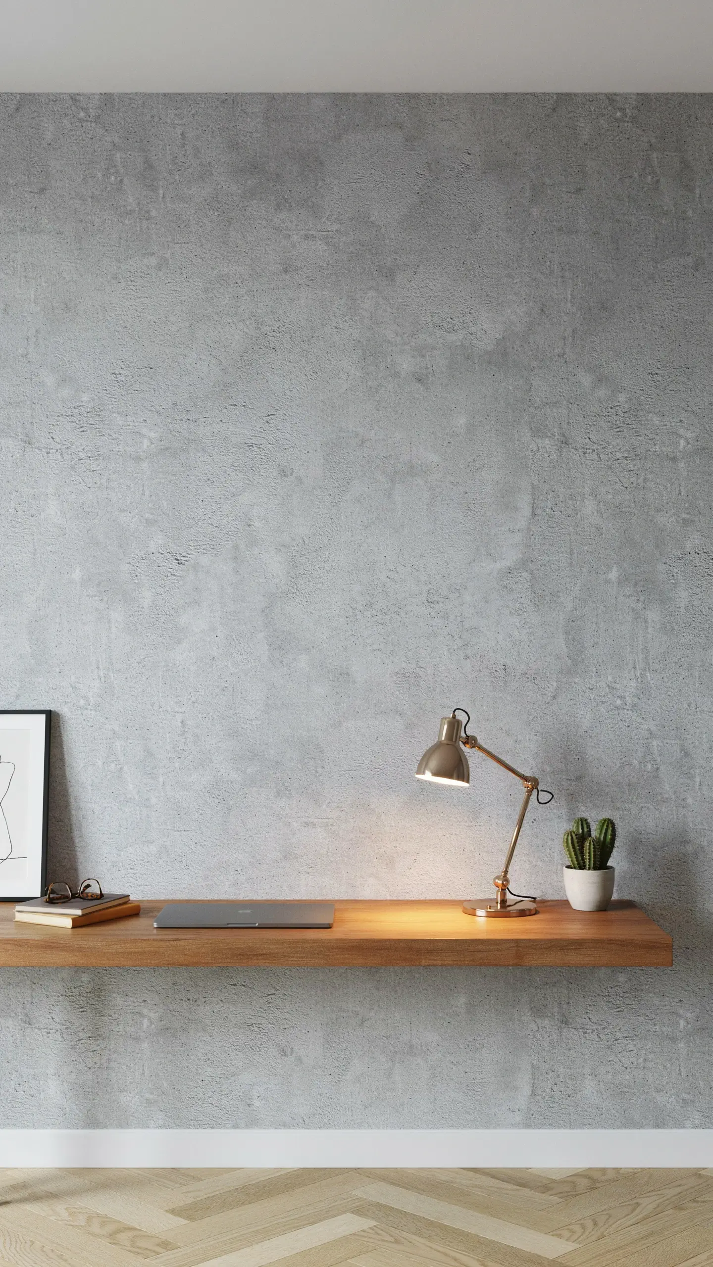 "A minimalist floating wooden desk workspace against a textured grey concrete or microcement accent wall, featuring a gold-toned brass desk lamp, a closed silver laptop, framed art, and a potted cactus." 
