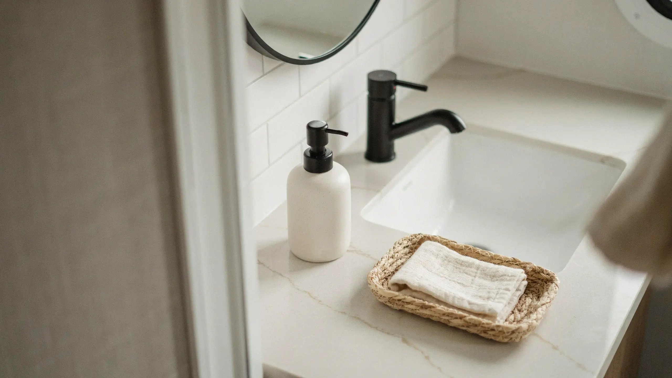 Minimalist bathroom counter styled with three items only: a matte black faucet, a white ceramic soap dispenser with a black pump, and a woven wicker tray holding a folded white towel on a marble vanity