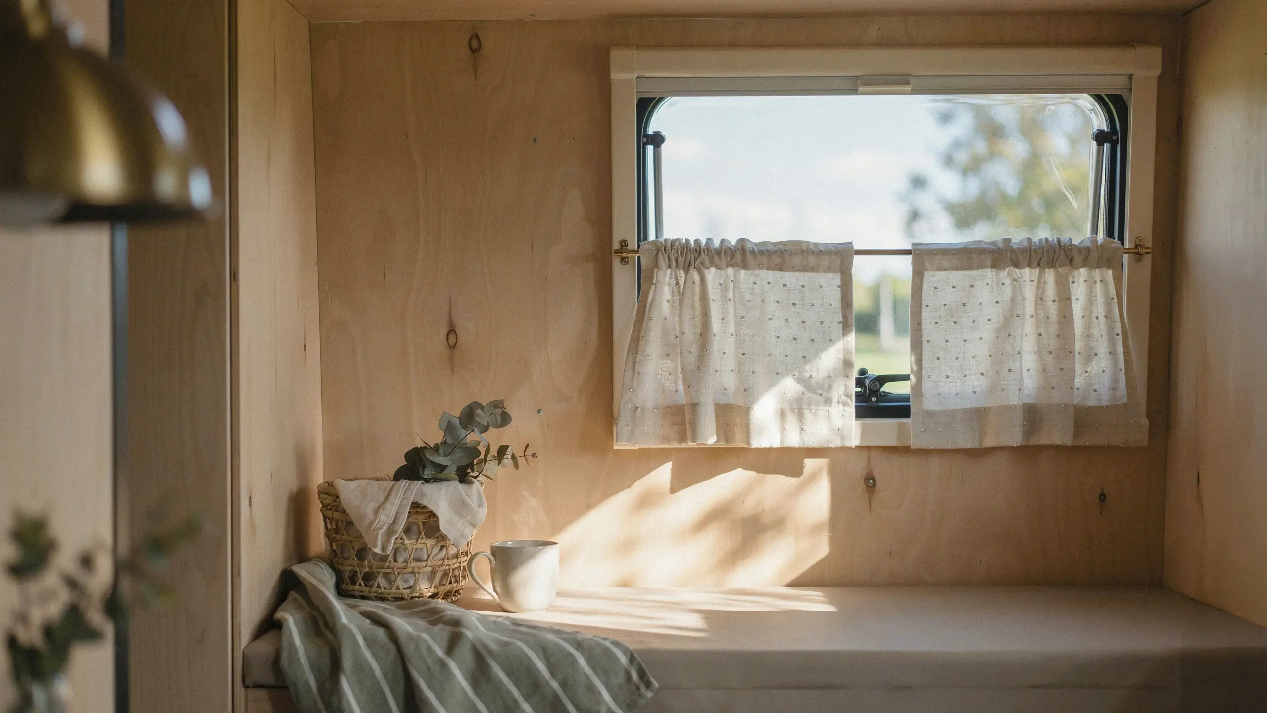 A bright, minimalist campervan interior featuring plywood walls and cafe curtains, with warm sunlight highlighting a small woven basket and mug, visually inspiring budget-friendly decor swaps like switching to a fresh duvet cover for the sleeping area