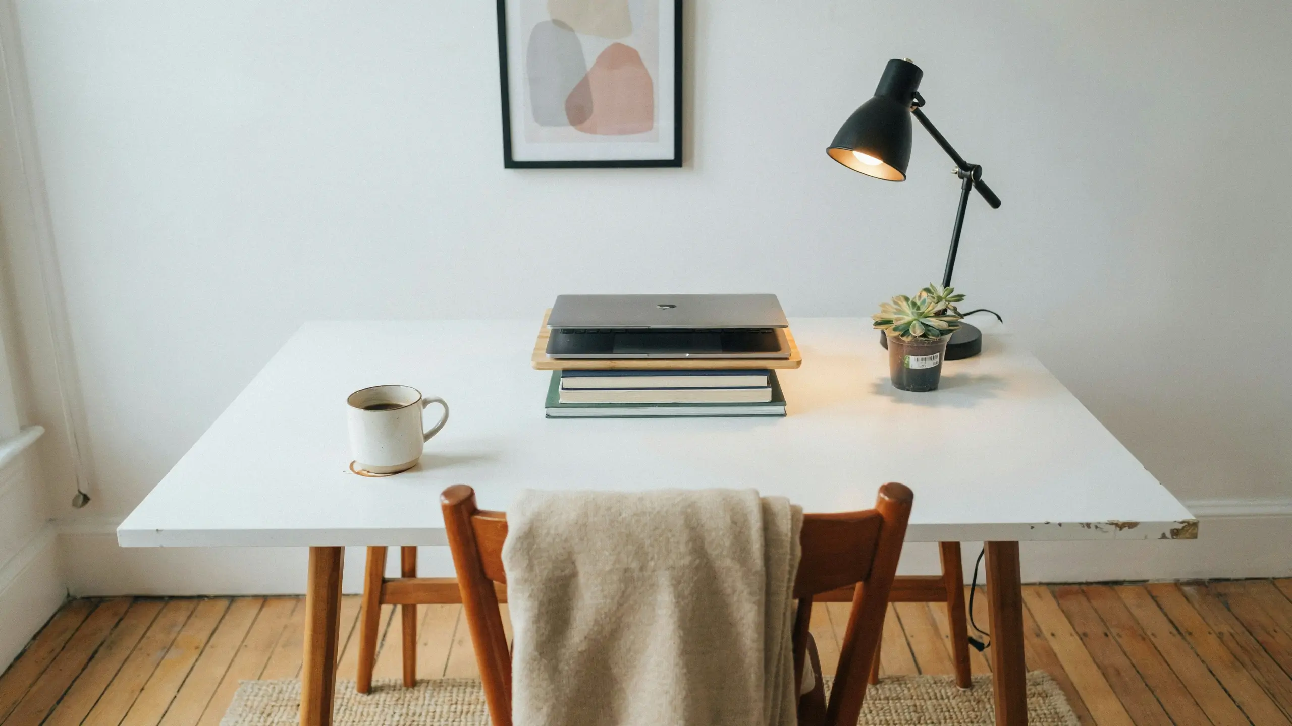 Minimalist white desk workspace with closed black laptop stacked on books, white coffee mug, black desk lamp, and small potted succulent for a cozy home office setting.