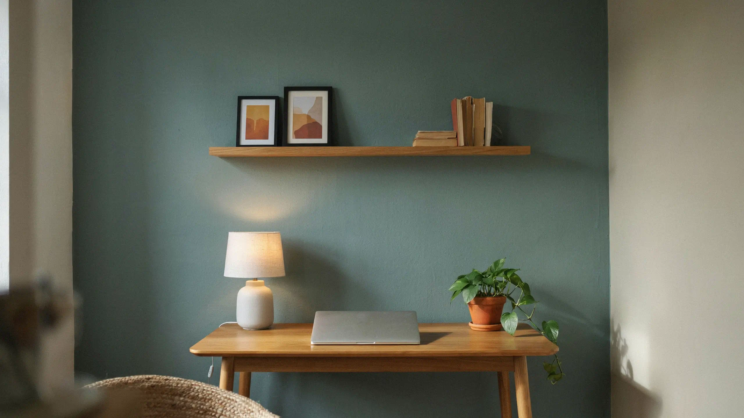 Minimalist wooden desk setup featuring a closed silver laptop, a warm-lit white table lamp, and a trailing pothos plant set against a calming teal accent wall with a floating shelf of books and framed art. 