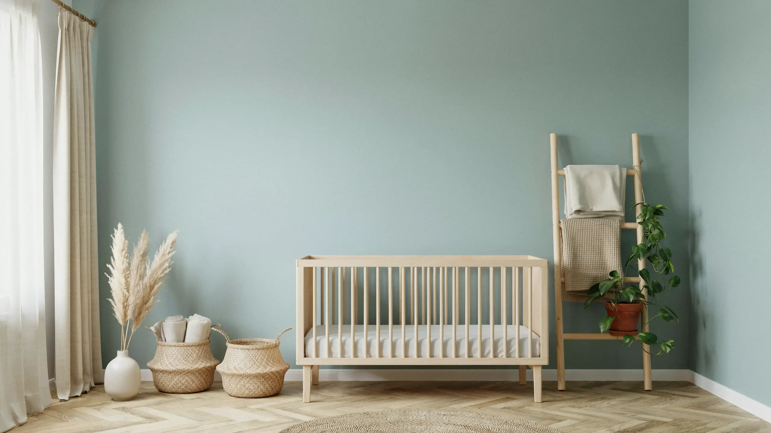 Minimalist light wood baby crib in a nursery with sage green walls, woven storage baskets, pampas grass, and a wooden blanket ladder with potted plant.