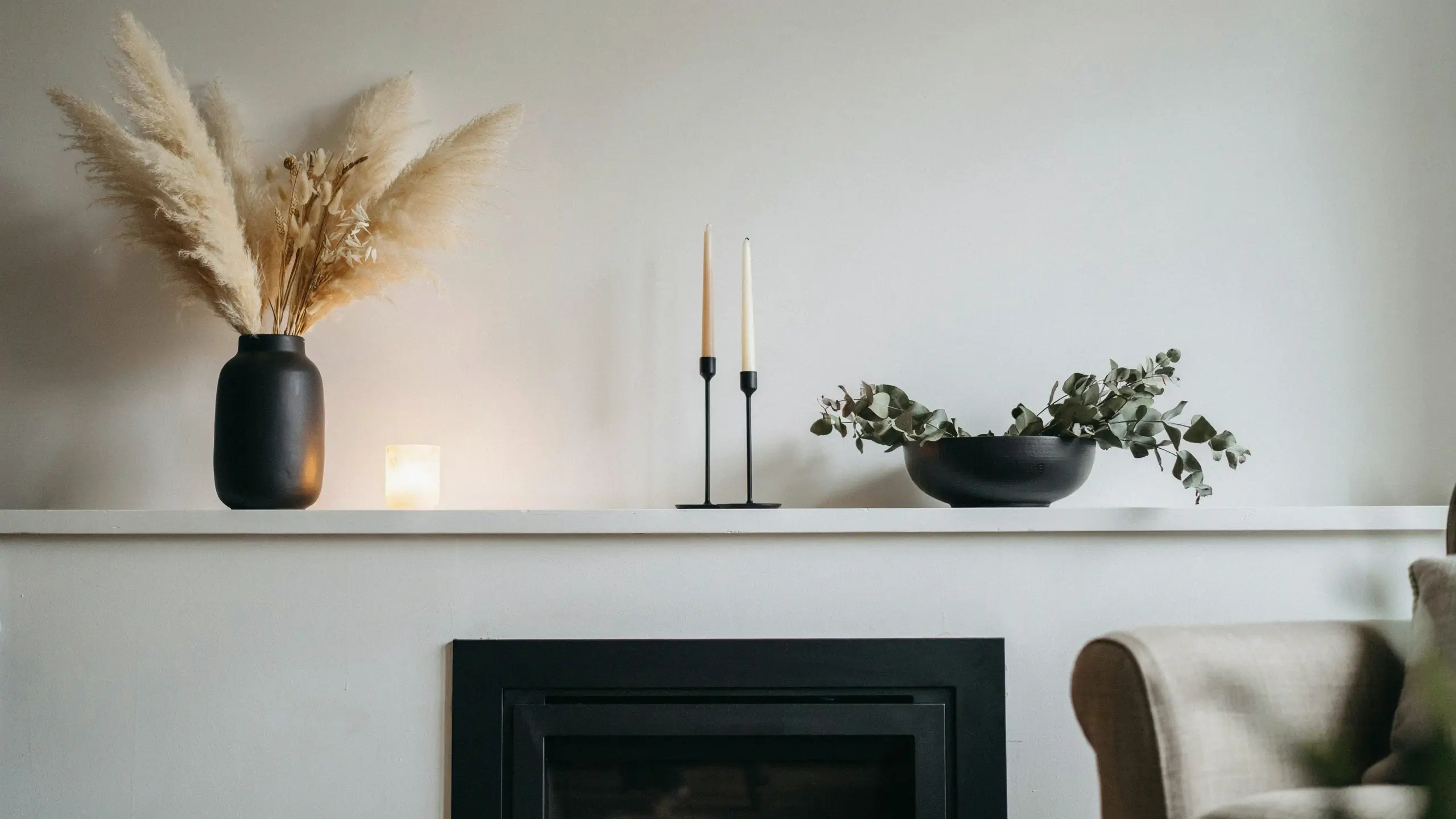 Minimalist fireplace mantel styled with a matte black vase filled with dried pampas grass, black candlesticks, glowing candle, and a bowl of eucalyptus leaves.