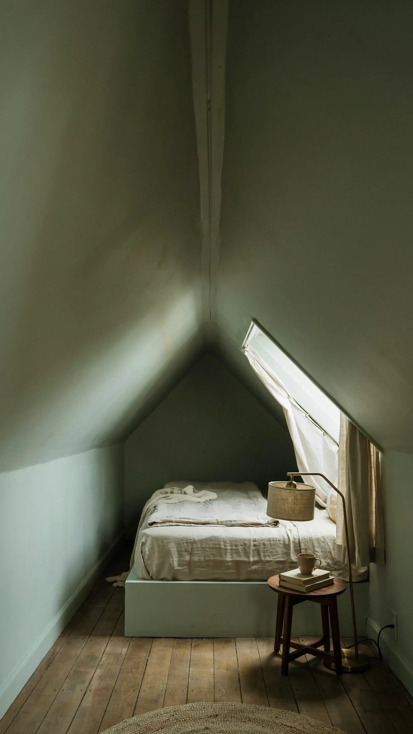 Minimalist sage green color-drenched attic bedroom with a skylight, white linen bedding, and a small wooden stool. 