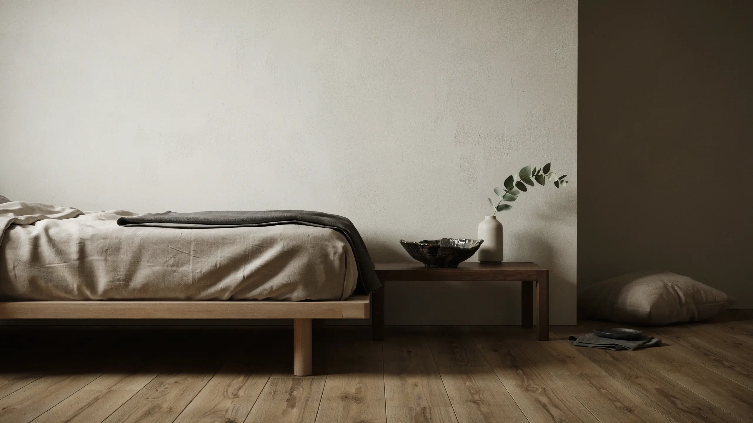 Minimalist Scandinavian bedroom design featuring a low natural wood platform bed with light beige linen sheets, a dark grey folded throw blanket, and a rustic wooden side table holding a white ceramic vase with eucalyptus branches and a dark ceramic bowl