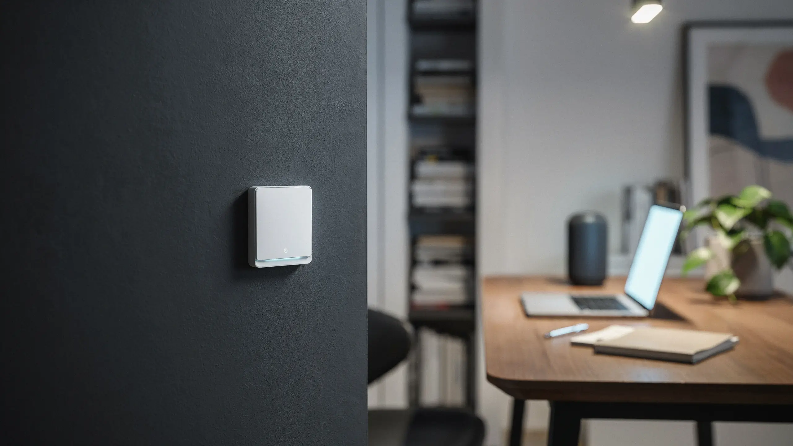 White minimalist smart touch switch with a subtle LED indicator mounted on a dark charcoal wall, positioned in a modern home office featuring a blurred background of a wooden desk and open laptop.