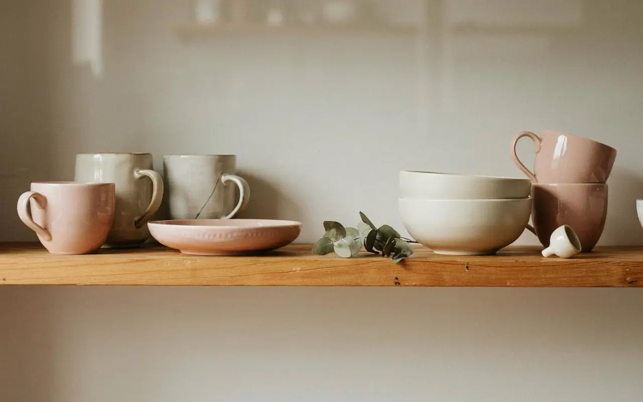 Mismatched cream and blush ceramic mugs, bowls, and plates grouped on a wooden open kitchen shelf.