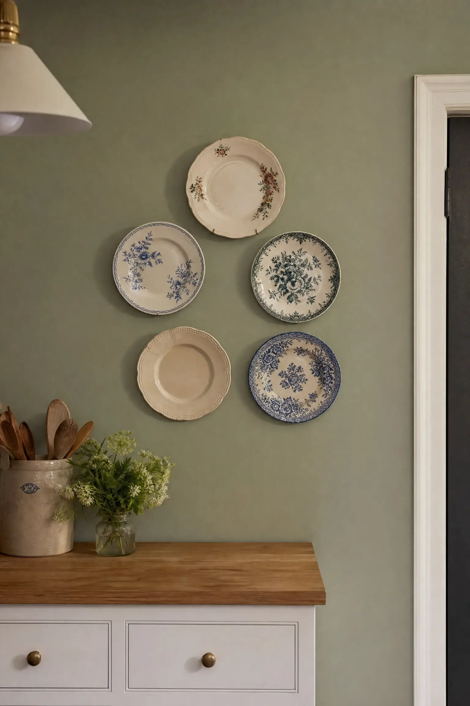 "Mismatched vintage floral and cream china plates hung in a loose cluster on a sage green kitchen wall above a wooden counter."