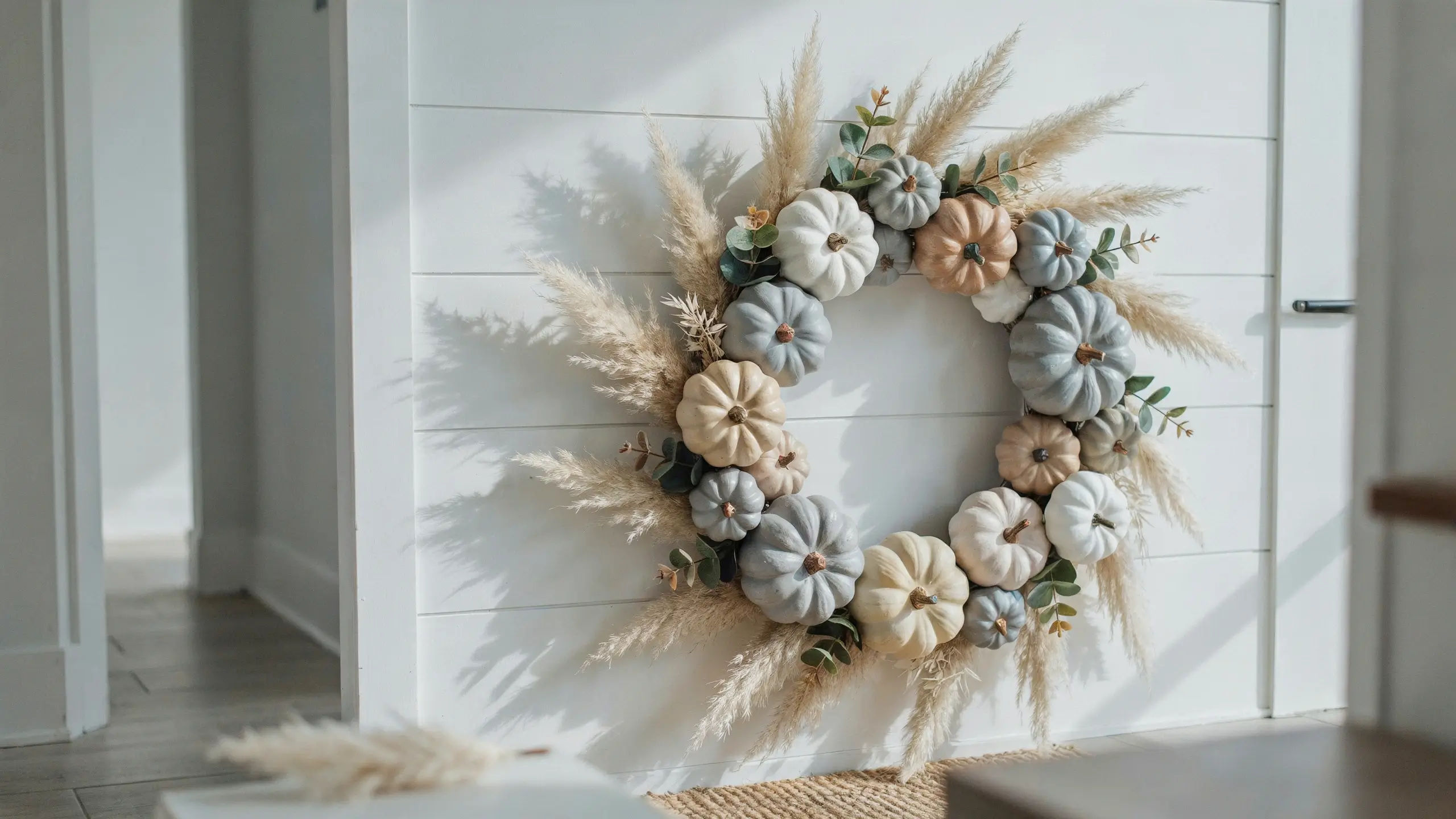 Modern farmhouse fall pumpkin wreath with white, grey, and cream fabric pumpkins, fluffy pampas grass, and eucalyptus leaves hanging against a white shiplap wall in a sunlit entryway.