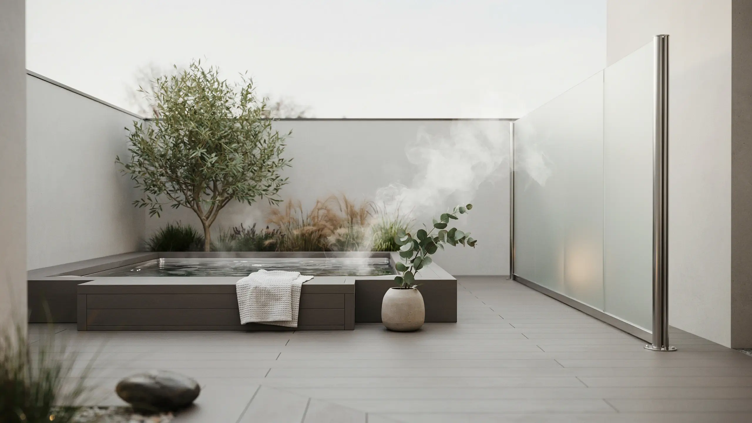 Modern outdoor hot tub with steamed water, enclosed by a sleek frosted glass or acrylic partial wall with stainless steel posts, styled alongside an olive tree and minimalist stone landscaping on a gray wood deck.