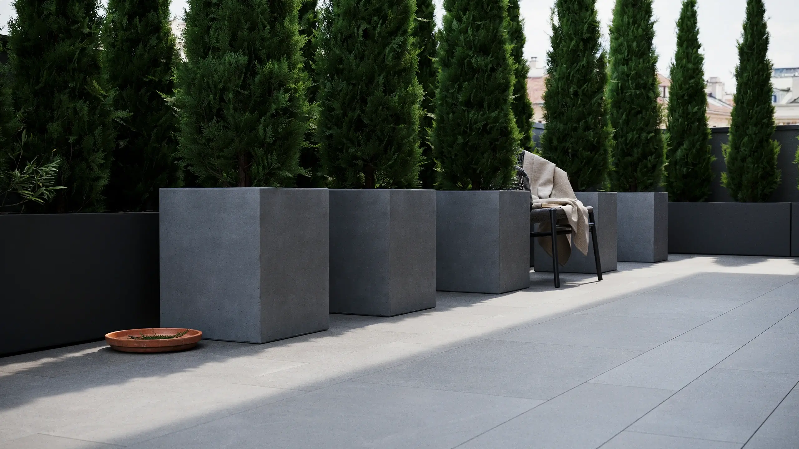 Tall grey square planters filled with dense green evergreen shrubs arranged along a modern outdoor patio with a black metal chair draped in a cream blanket.