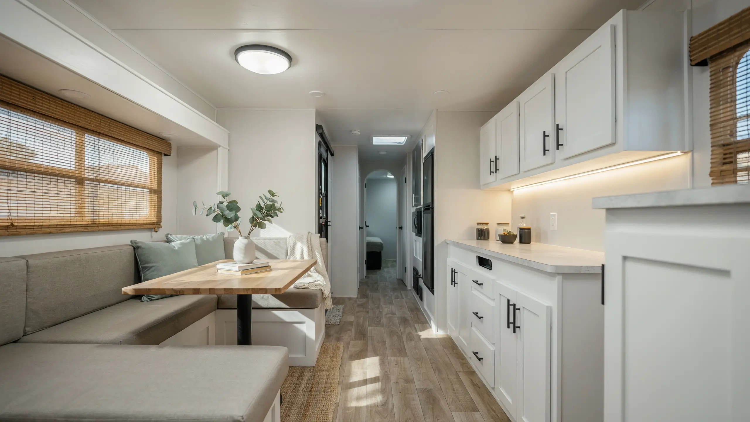 Modern farmhouse RV interior renovation featuring a white shaker kitchen with matte black hardware and a light grey dinette booth paired with a butcher block table and sage green throw pillows. 