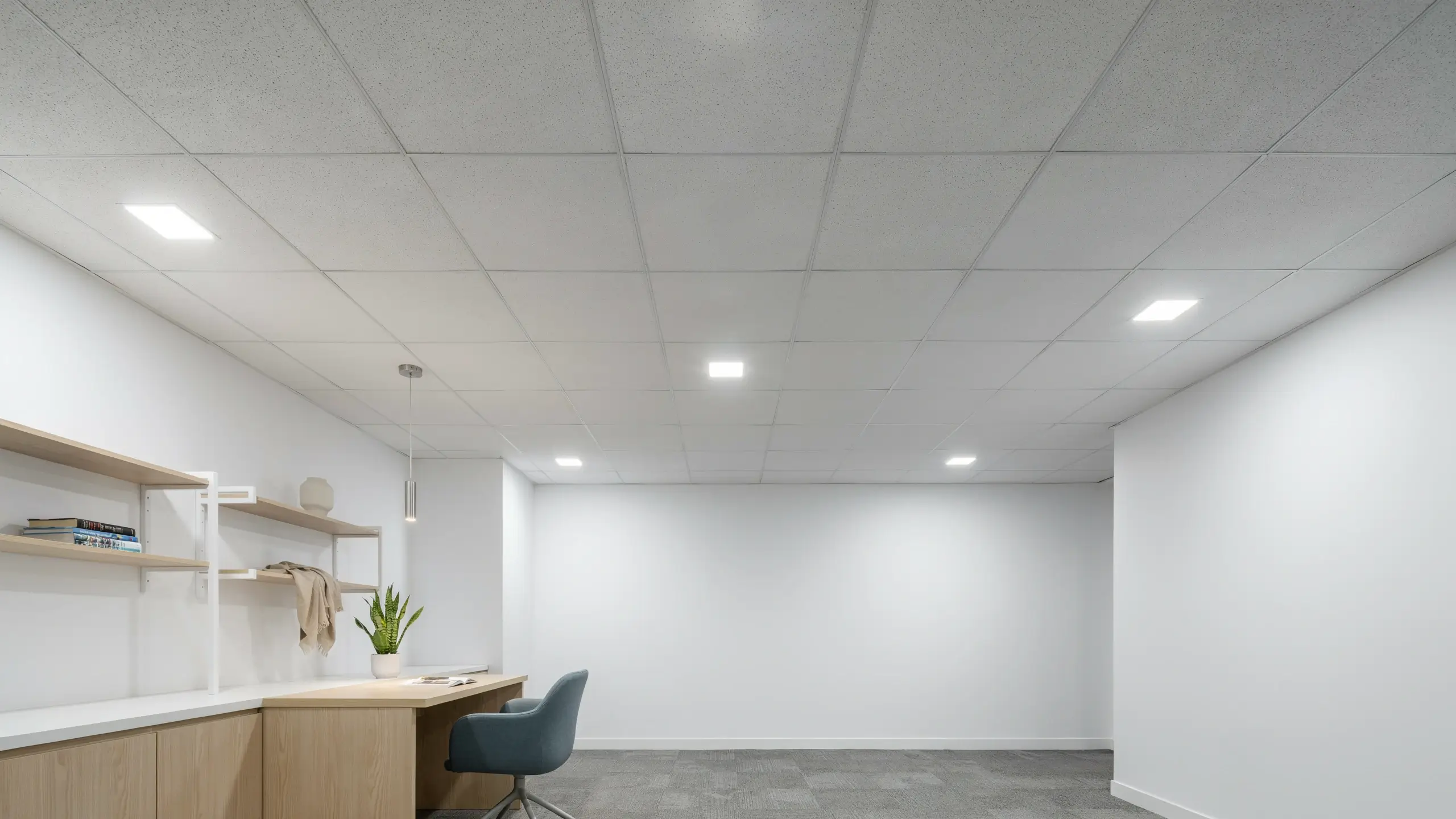 Modern suspended drop ceiling with white textured tiles and square LED panel lights illuminating a clean, minimalist office workspace with light wood built-in desk and blue swivel chair.