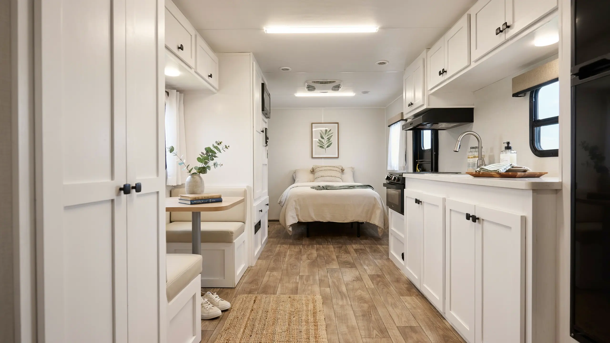 Modern white RV interior remodel showcasing a bright open-concept kitchen with white shaker cabinets, matte black hardware, a dining booth, and a cozy rear bedroom decorated with neutral linens and botanical wall art. 
