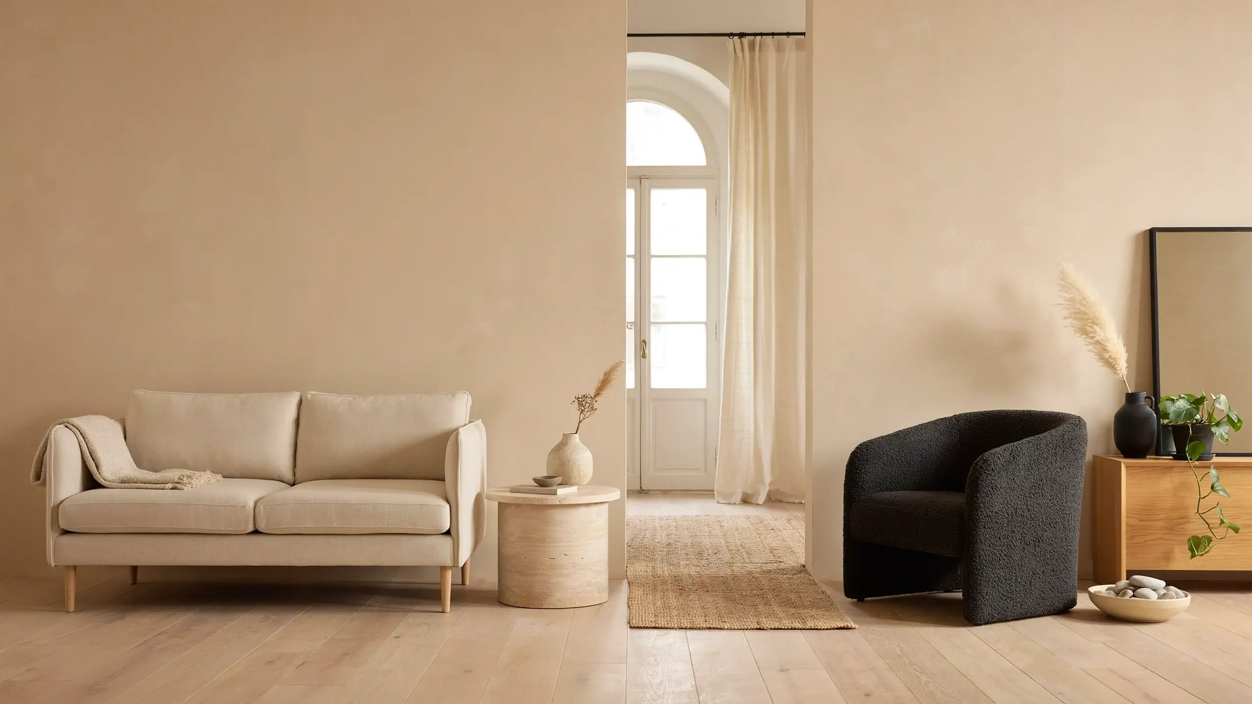 Minimalist monochromatic beige living room with a light cream sofa, round wooden side table holding a pampas grass vase, textured charcoal black armchair, wooden console table with a black-framed mirror and potted plant, illuminated by natural light from a white arched glass doorway.