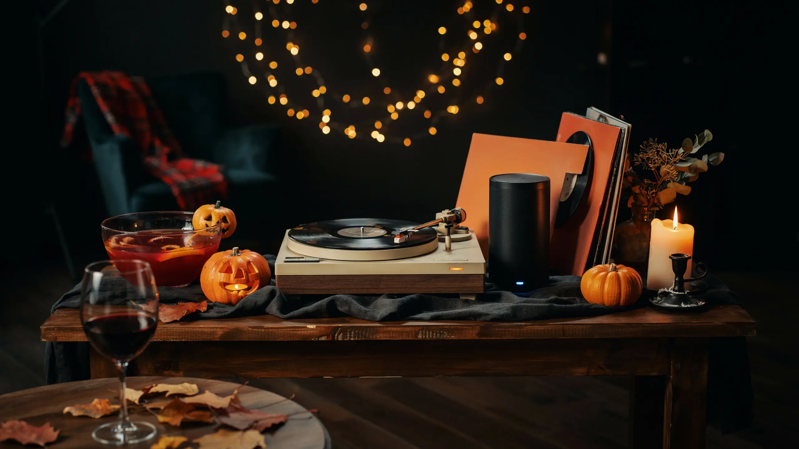 "Cozy autumn tabletop setup featuring a white vintage record player, black smart speaker, orange vinyl, a glass of wine, and glowing carved pumpkins under warm fairy lights for a moody Halloween music evening."