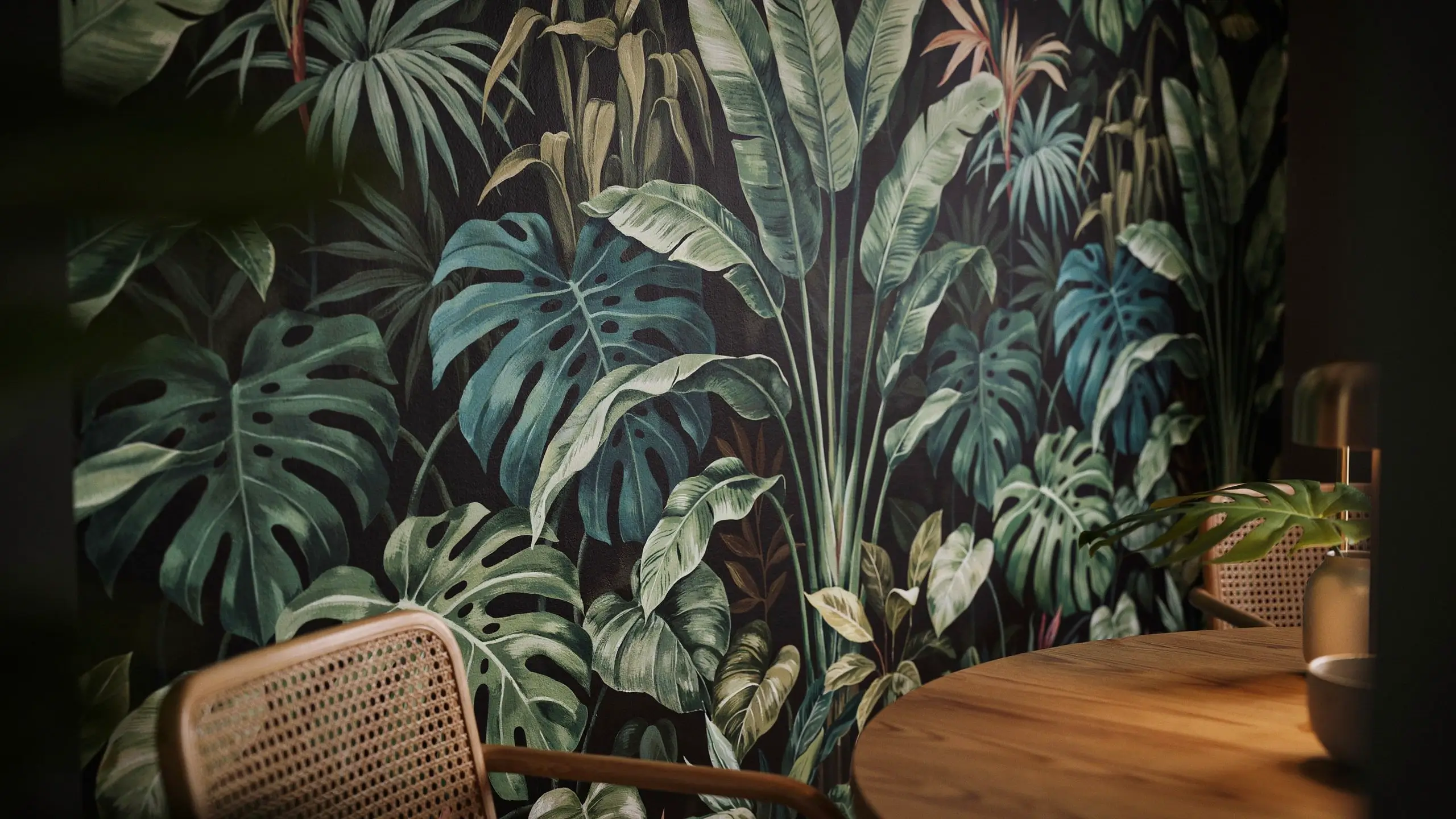 Moody dining corner featuring a dramatic large-scale botanical wallpaper with oversized monstera and palm leaves, paired with a rattan chair and wooden table. 