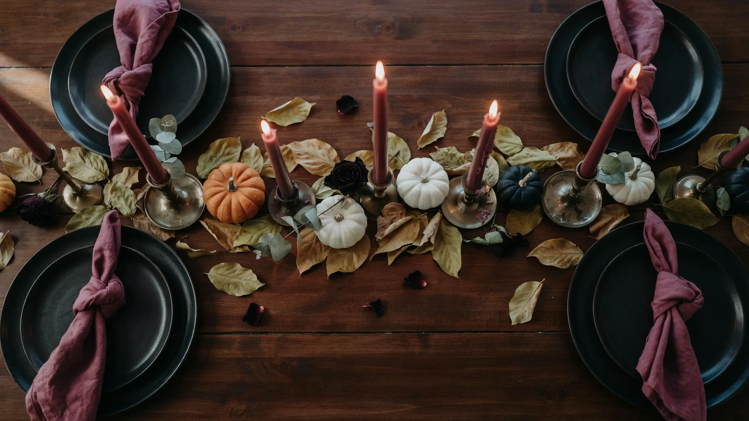 Flat lay of dark autumnal table setting with matte black plates, dusty rose knotted napkins, lit burgundy taper candles, and small white, orange, and dark green pumpkins on a rustic wood table. 