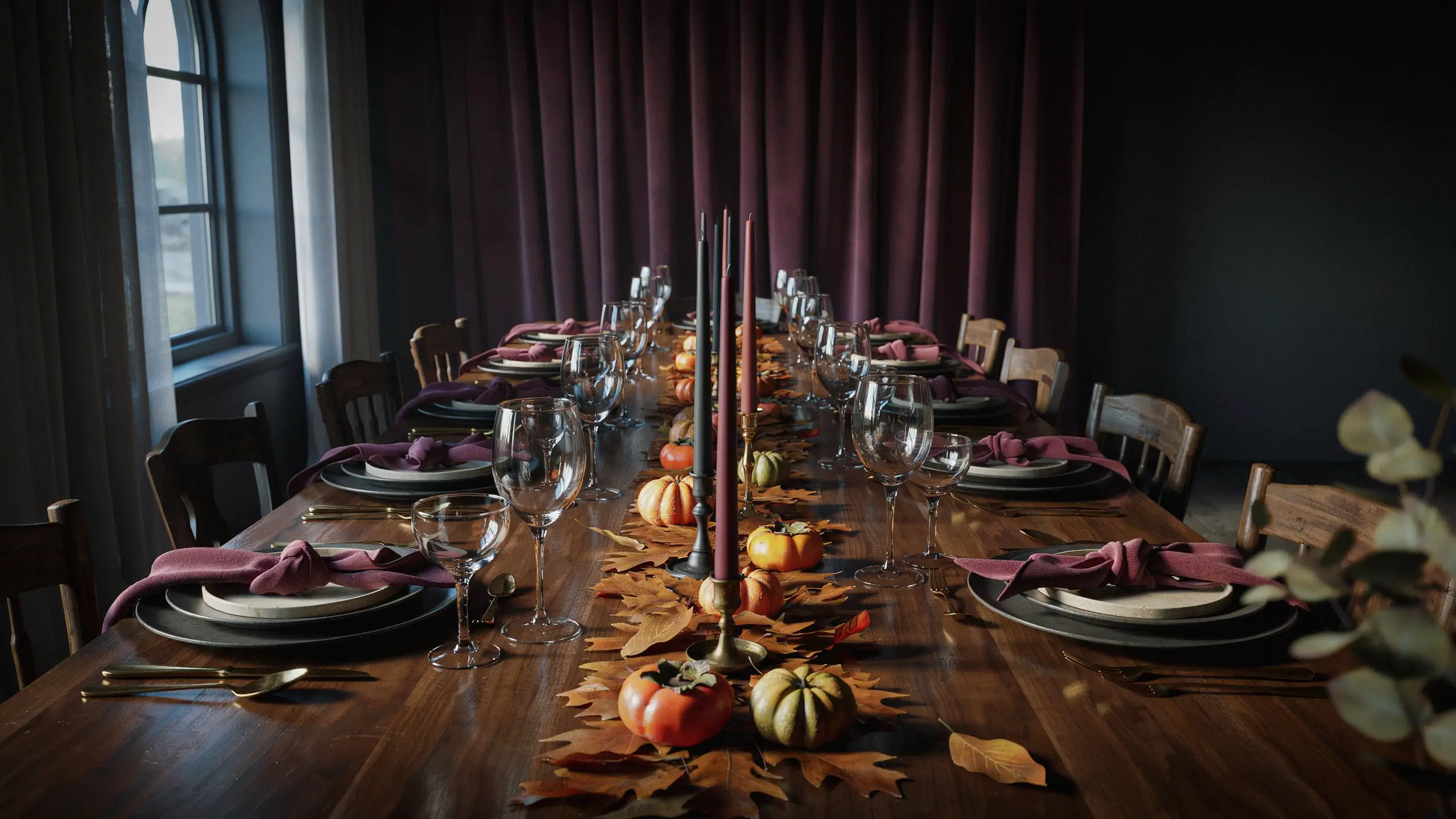 Long wooden dining table arranged for an autumn feast with burgundy napkins, gold cutlery, tall taper candles, and mini pumpkins against a dark plum curtain backdrop. 