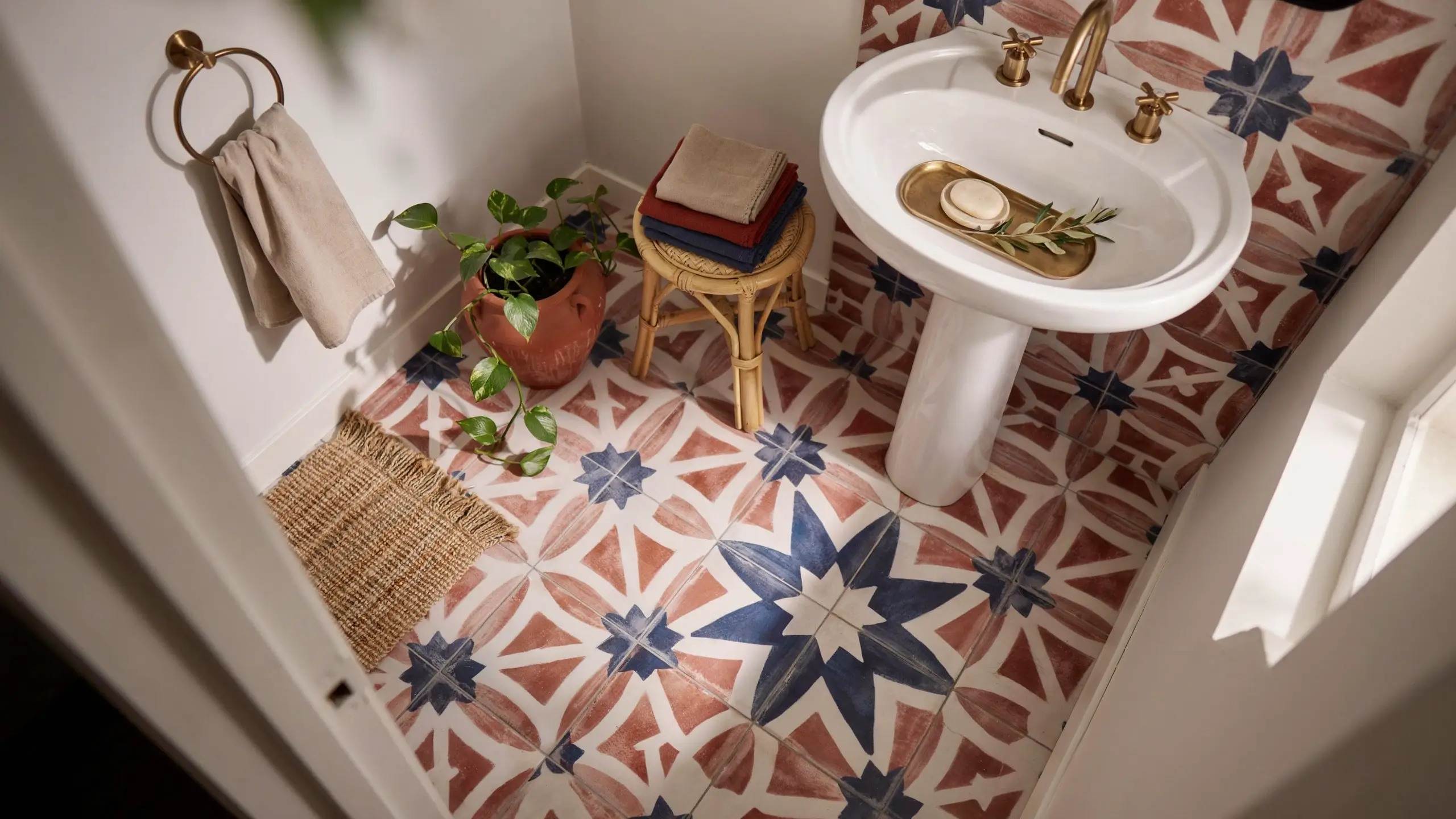 Close-up view of a bathroom featuring a Moroccan-inspired geometric tile floor with terracotta and navy blue starburst patterns, surrounded by a white pedestal sink with brass fixtures, a woven rattan stool, and a potted pothos plant