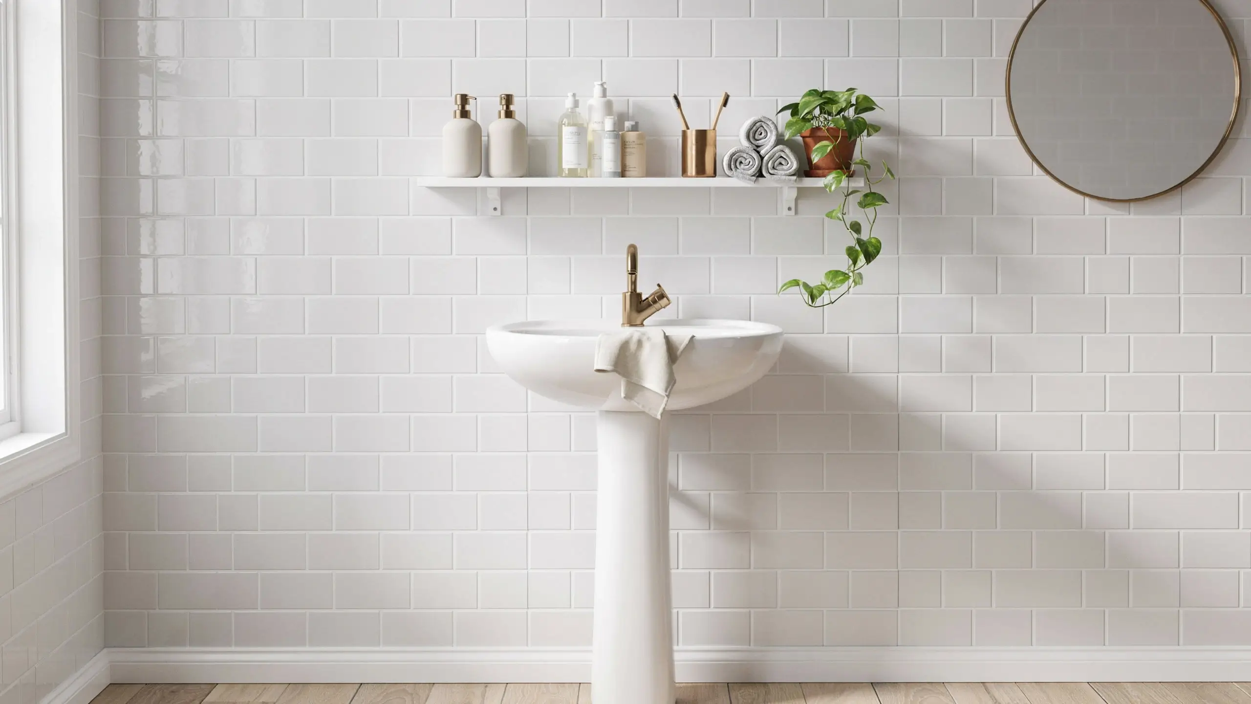 White MOSSLANDA picture ledge holding toiletries and a pothos plant over a white pedestal sink with a brass faucet in a modern bathroom with subway tiles and a round mirror.