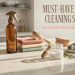 Flat lay of must-have kitchen cleaning supplies including an amber glass spray bottle, wooden scrub brushes, baking soda, and reusable cloths on a marble counter.