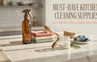 Flat lay of must-have kitchen cleaning supplies including an amber glass spray bottle, wooden scrub brushes, baking soda, and reusable cloths on a marble counter.