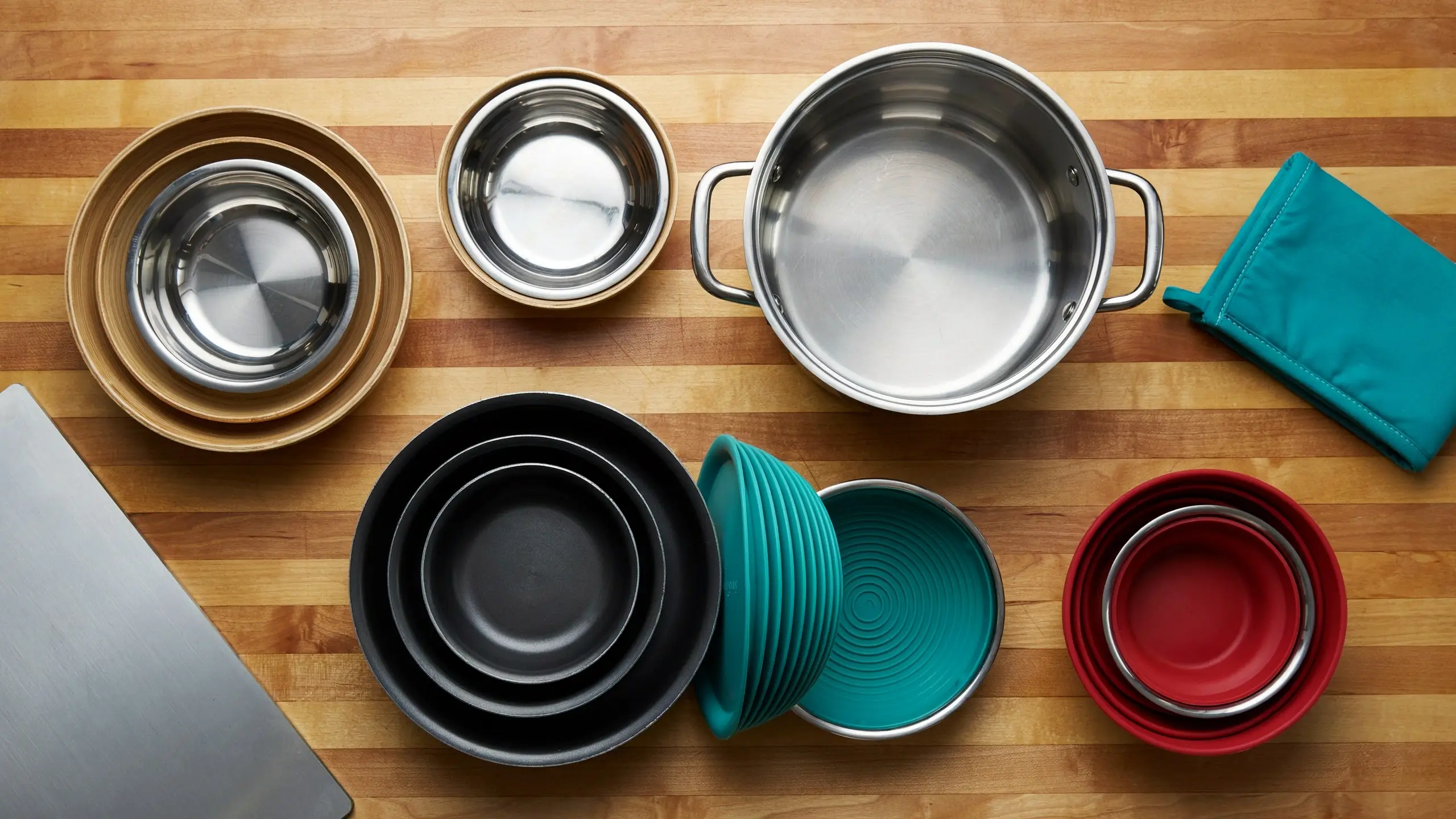 Top-down view of a space-saving nesting cookware set featuring stainless steel pots, black and red mixing bowls, and a teal collapsible silicone lid arranged on a wooden kitchen countertop. 