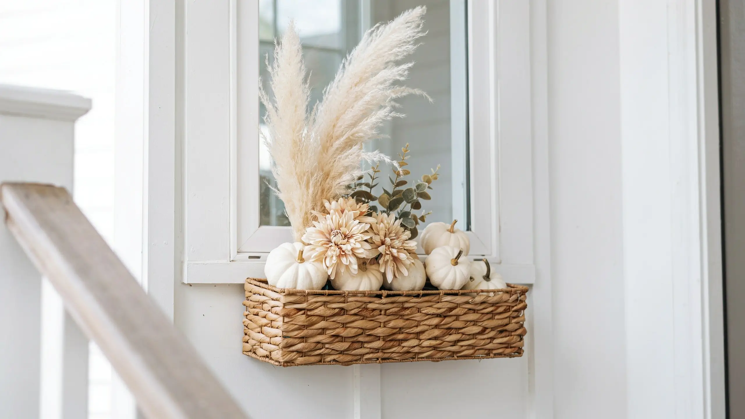 "A textured woven wicker basket mounted on a white front door, styled with white mini pumpkins, cream dahlias, dried eucalyptus leaves, and tall feathery pampas grass for a minimalist seasonal entryway." 