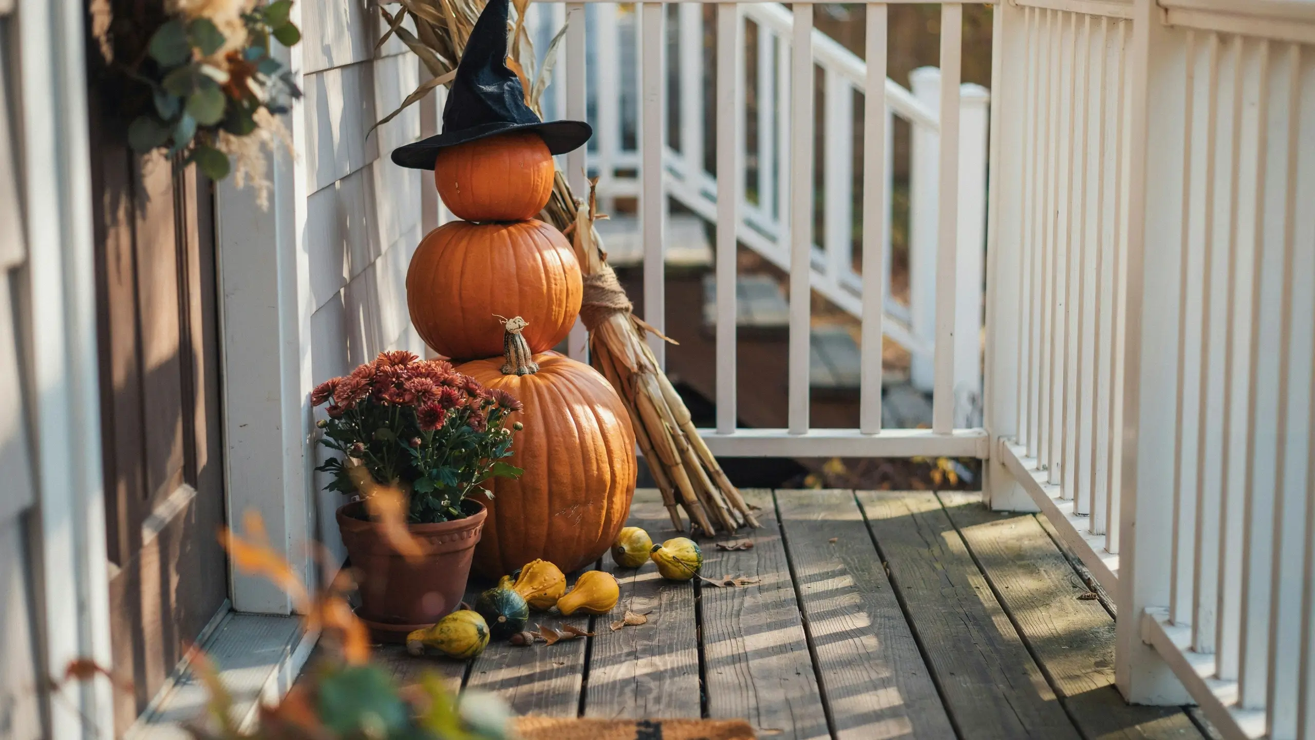 A rustic fall porch decor idea featuring a no-carve stacked pumpkin tower topped with a black witch hat, sitting next to a potted red chrysanthemum, scattered yellow gourds, and tied dried corn stalks on a sunny wooden deck.