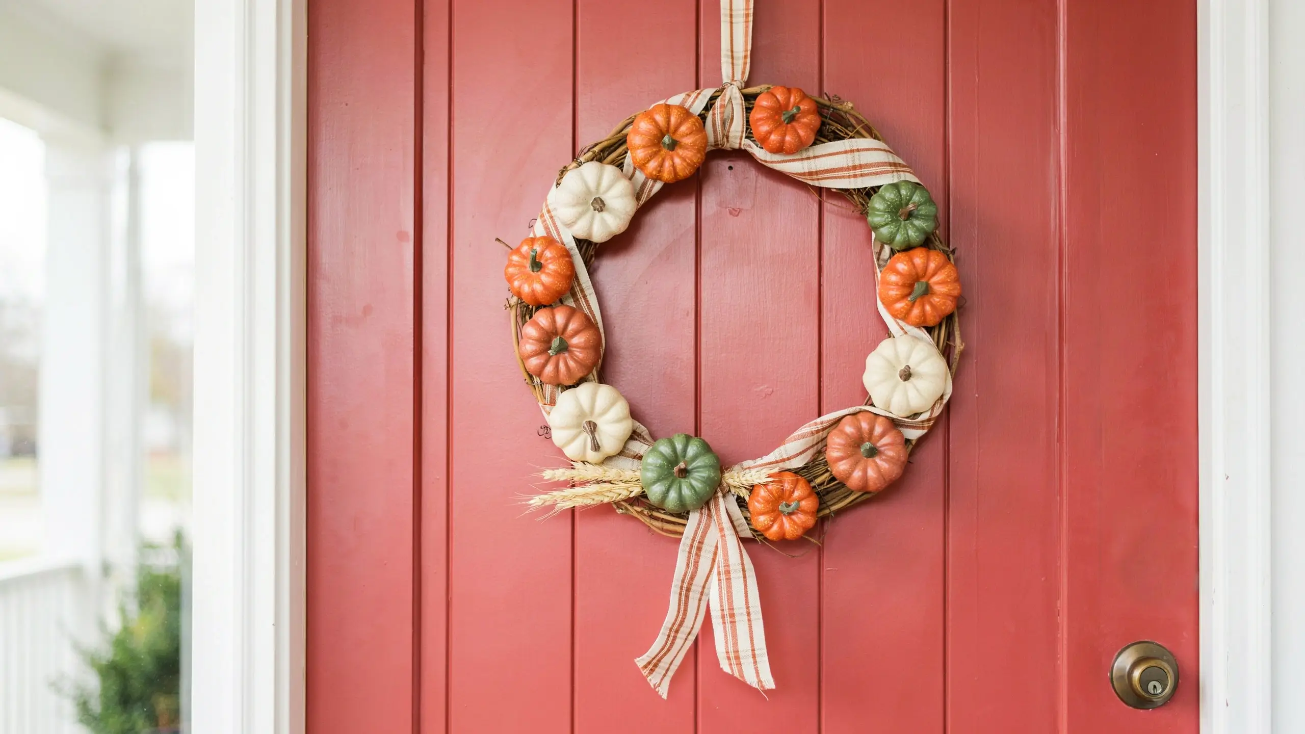 A rustic no-carve fall wreath featuring a grapevine base decorated with mini orange, green, and white pumpkins and a white-and-orange plaid ribbon, hanging on a red paneled front door for farmhouse autumn porch decor.