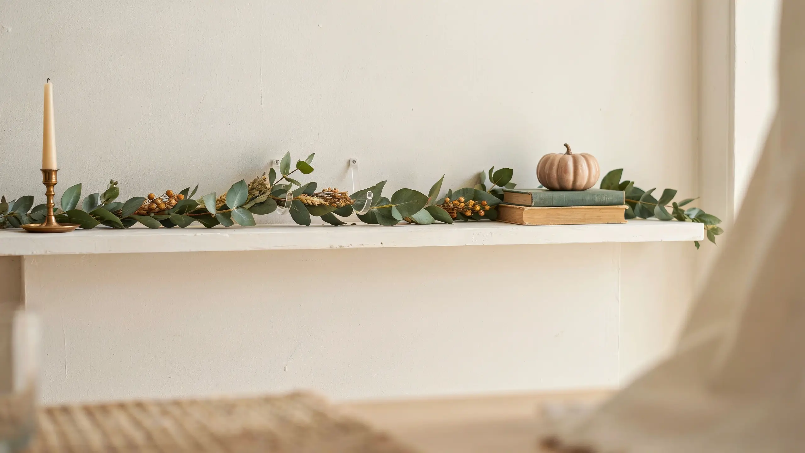 Rental-friendly fall shelf decor featuring beige pumpkin on stacked books, candlestick, and eucalyptus garland hanging with no-drill clear adhesive hooks.