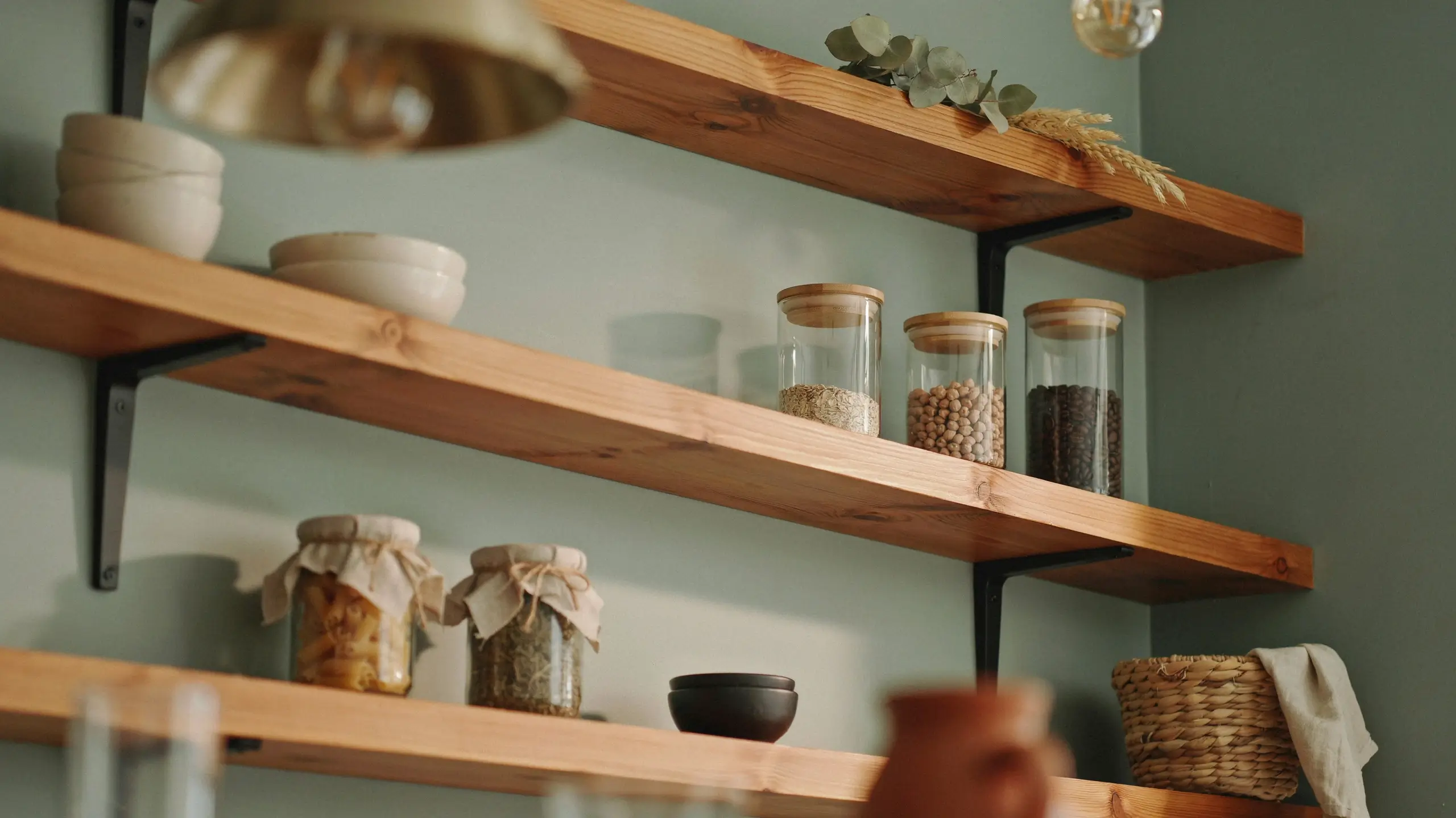 Open wood shelving with glass pantry jars and ceramic bowls on a sage green wall. 