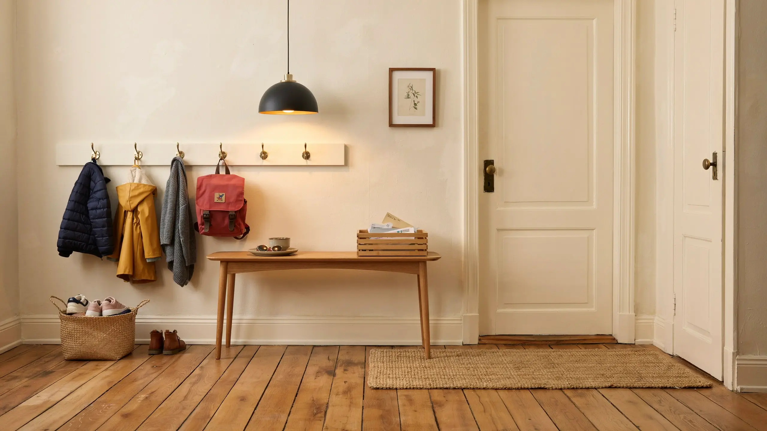 A well-lit minimalist entryway featuring a wooden bench, a white coat rack with a blue jacket, yellow jacket, and red backpack, alongside a wicker basket of shoes and a slatted crate holding mail by the front door