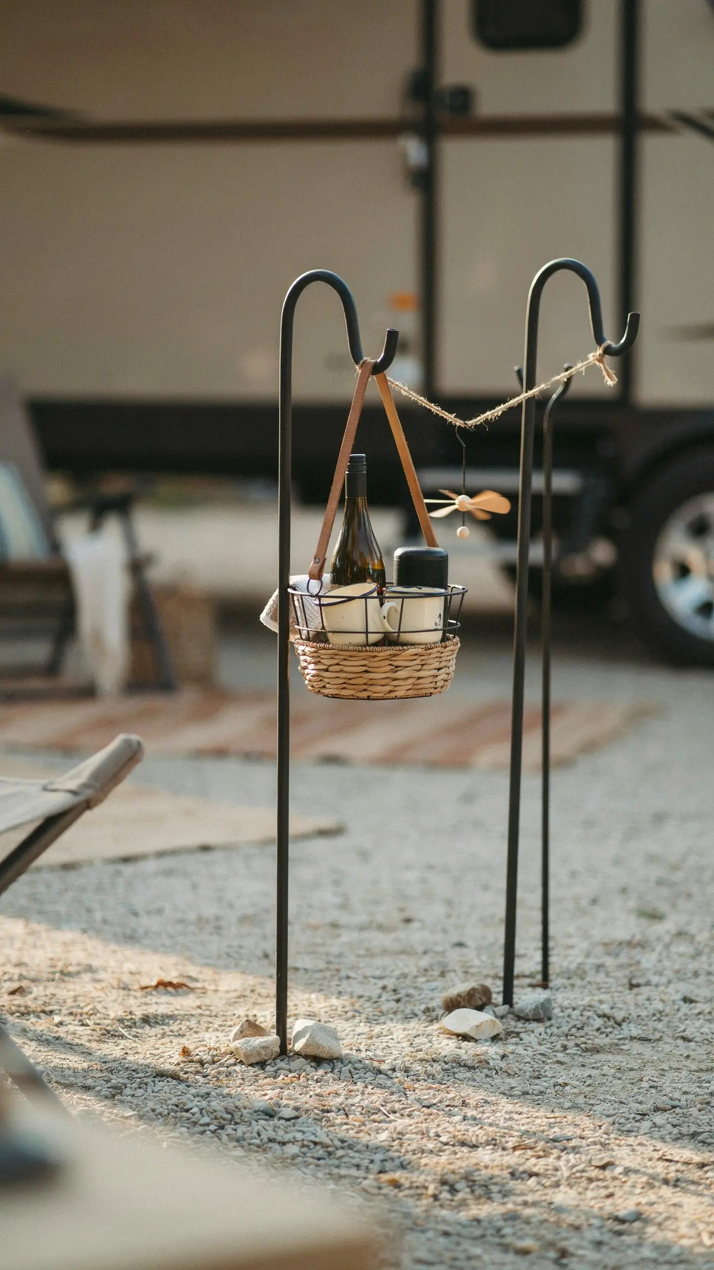 Rustic hanging bar cart made from a woven basket and leather straps suspended on tall black shepherd's hooks, holding a wine bottle, white mug, and travel cup on a gravel campsite beside an RV. 