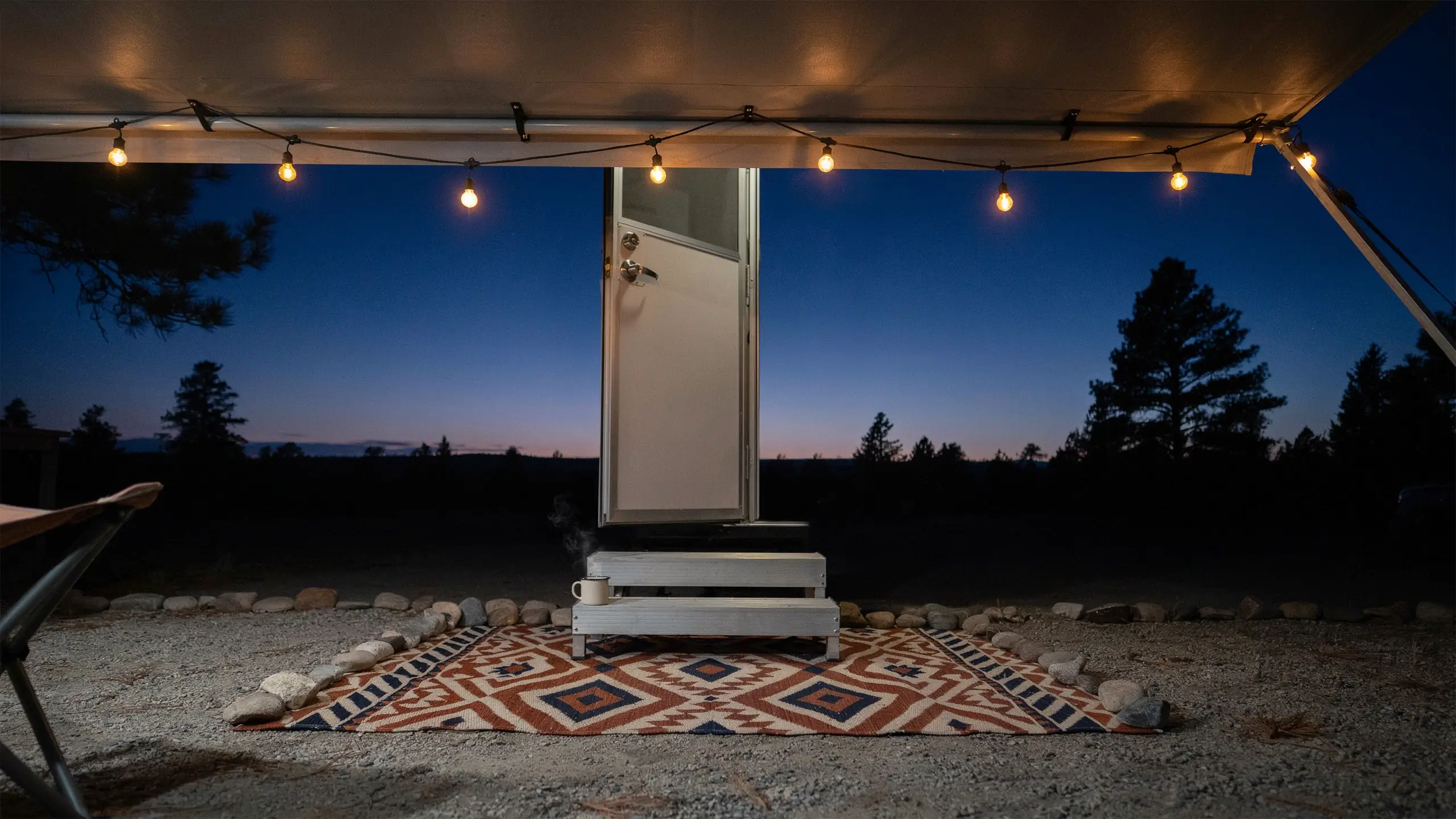 Outdoor rug and string lights at the camper entry, with a geometric mat and a steaming mug on the stairs against a twilight sky