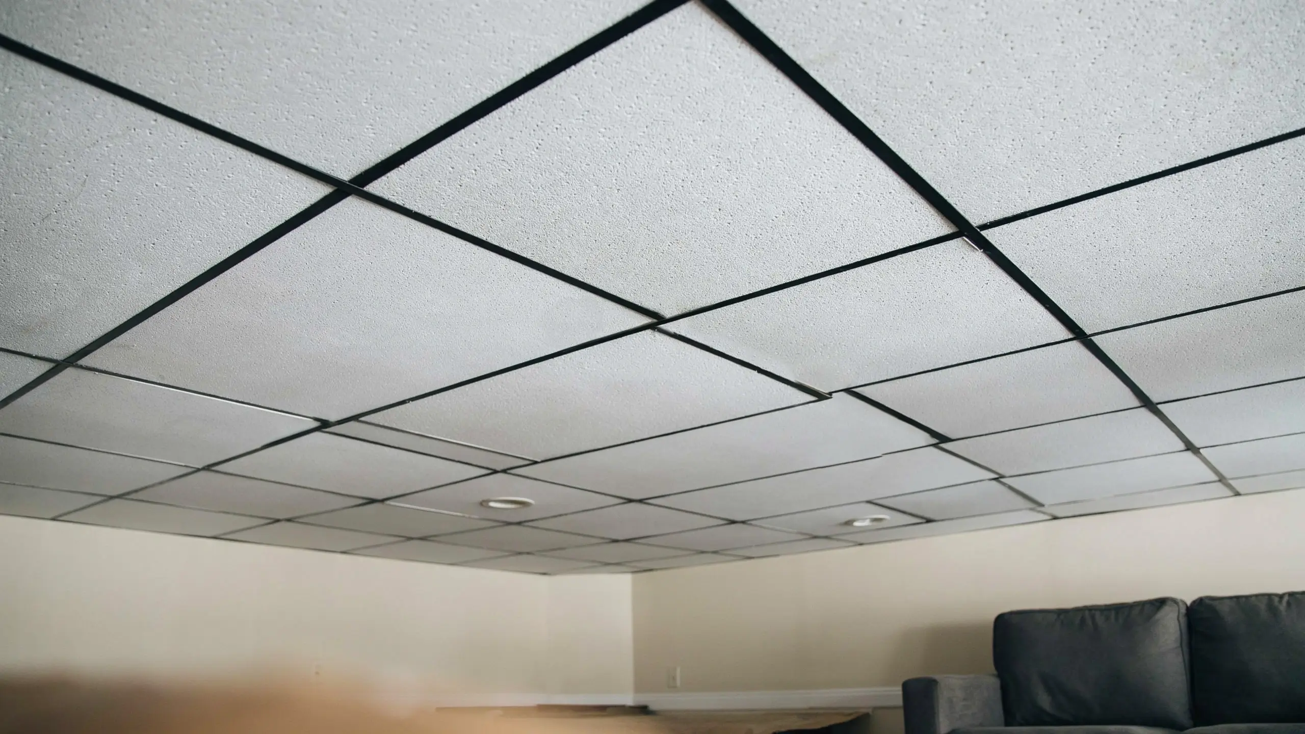 White acoustic tiles and a painted black metal drop ceiling grid in a finished basement with beige walls.