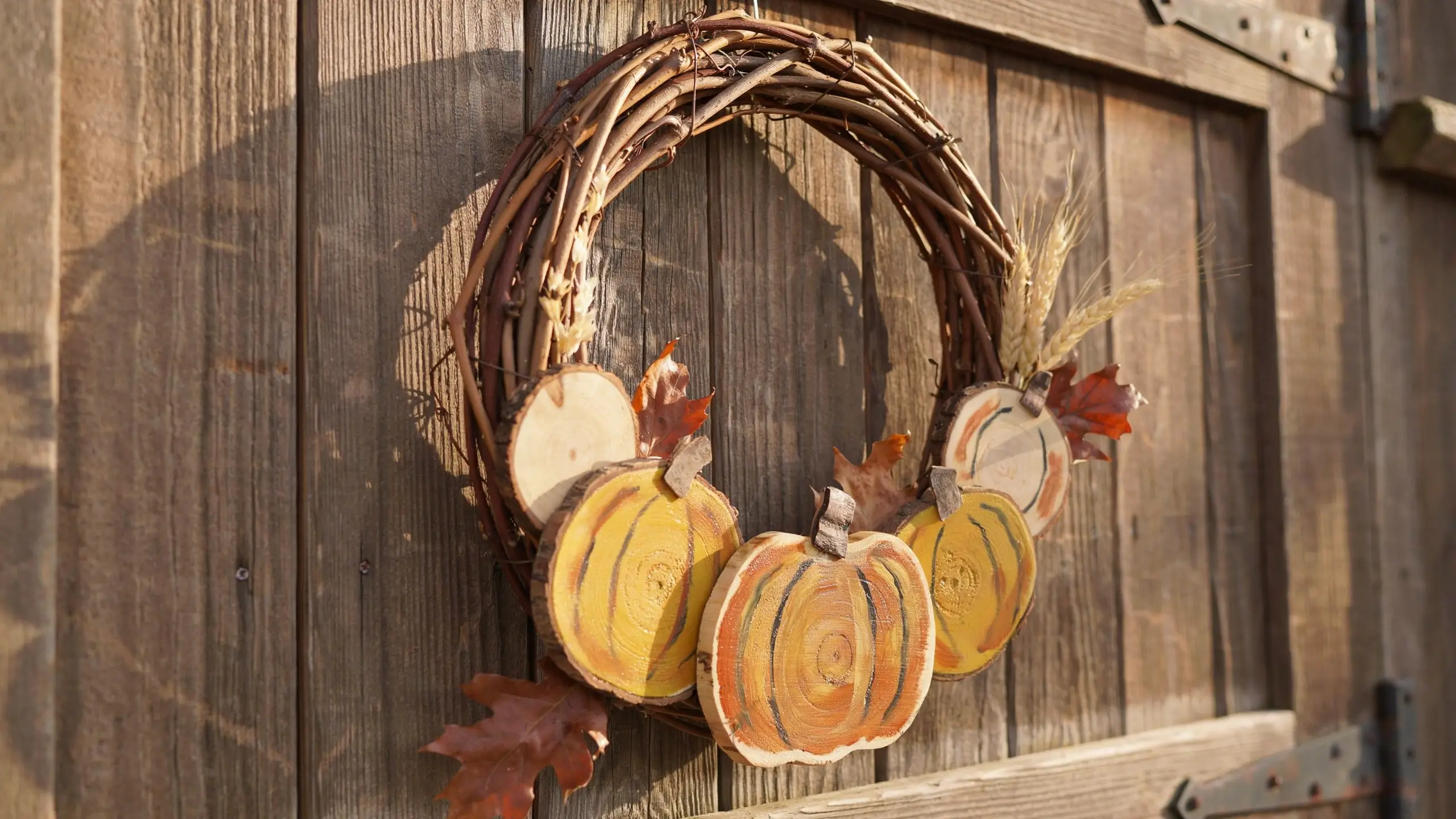 Rustic grapevine wreath featuring hand-painted wood slice pumpkins, dried wheat stalks, and brown autumn leaves hanging on a weathered wooden barn door.