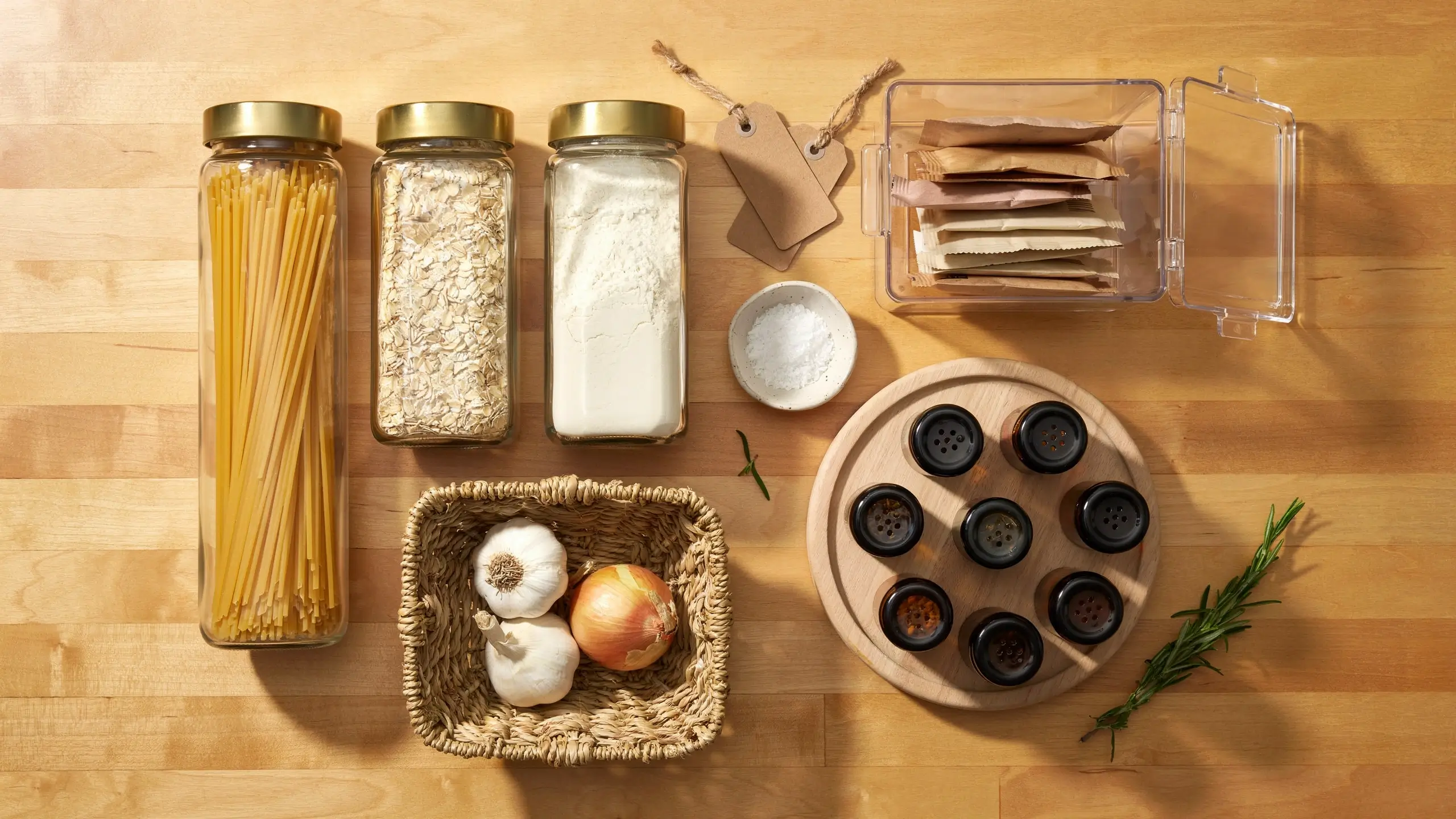 Overhead view of organized pantry storage solutions including glass jars with brass lids for pasta, oats, and flour, a wooden spice rack with small round containers, a woven basket holding fresh garlic and onion, and a clear acrylic bin containing tea bags, all laid out on a light wood kitchen surface. 