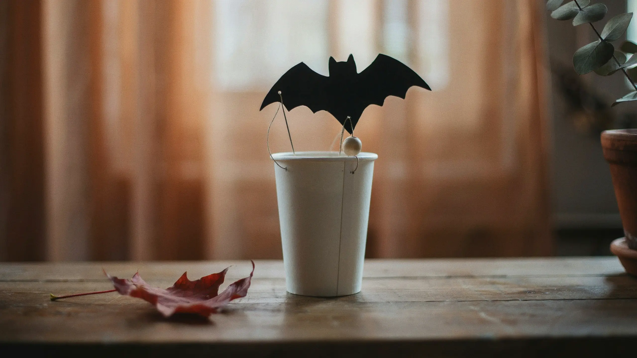DIY flying bat craft made from a white paper cup and black construction paper, suspended by wire on a rustic wooden table beside a red autumn leaf.