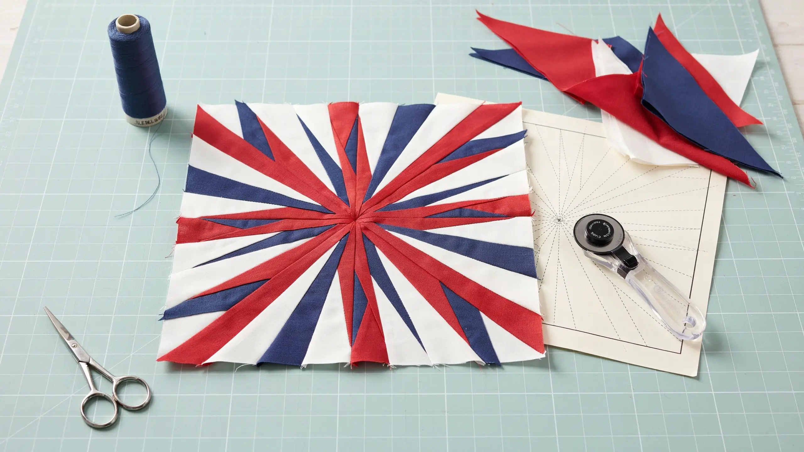 Finished red, white, and blue foundation paper pieced fireworks quilt block resting on a light green gridded cutting mat, surrounded by a spool of navy thread, silver sewing scissors, a rotary cutter, and fabric scraps.