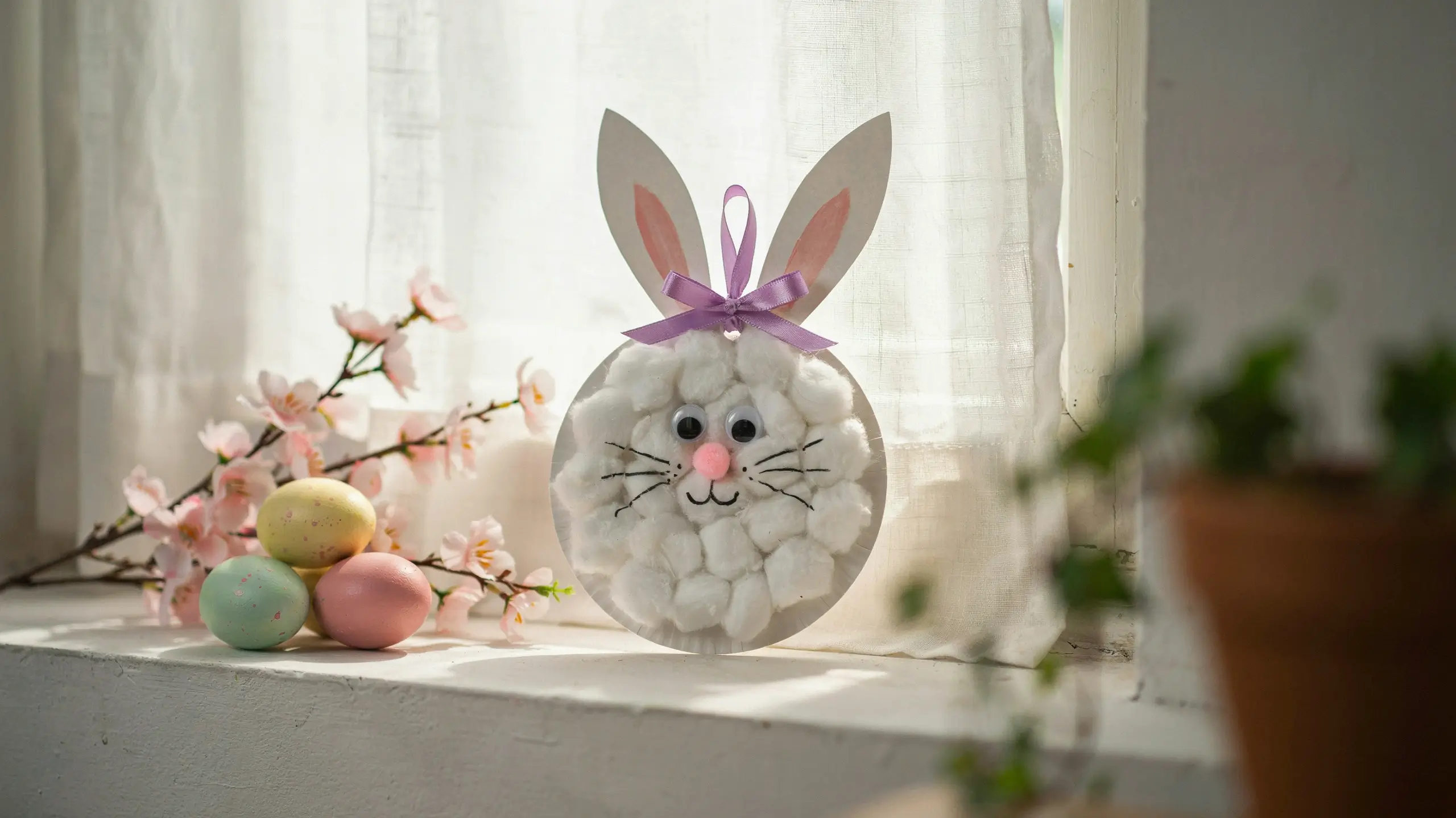 A close-up photo of a DIY paper plate bunny craft decorated with fluffy white cotton balls, googly eyes, a pink pom-pom nose, and a purple satin ribbon bow, sitting on a sunny windowsill next to pastel Easter eggs and pink spring cherry blossom branches. 