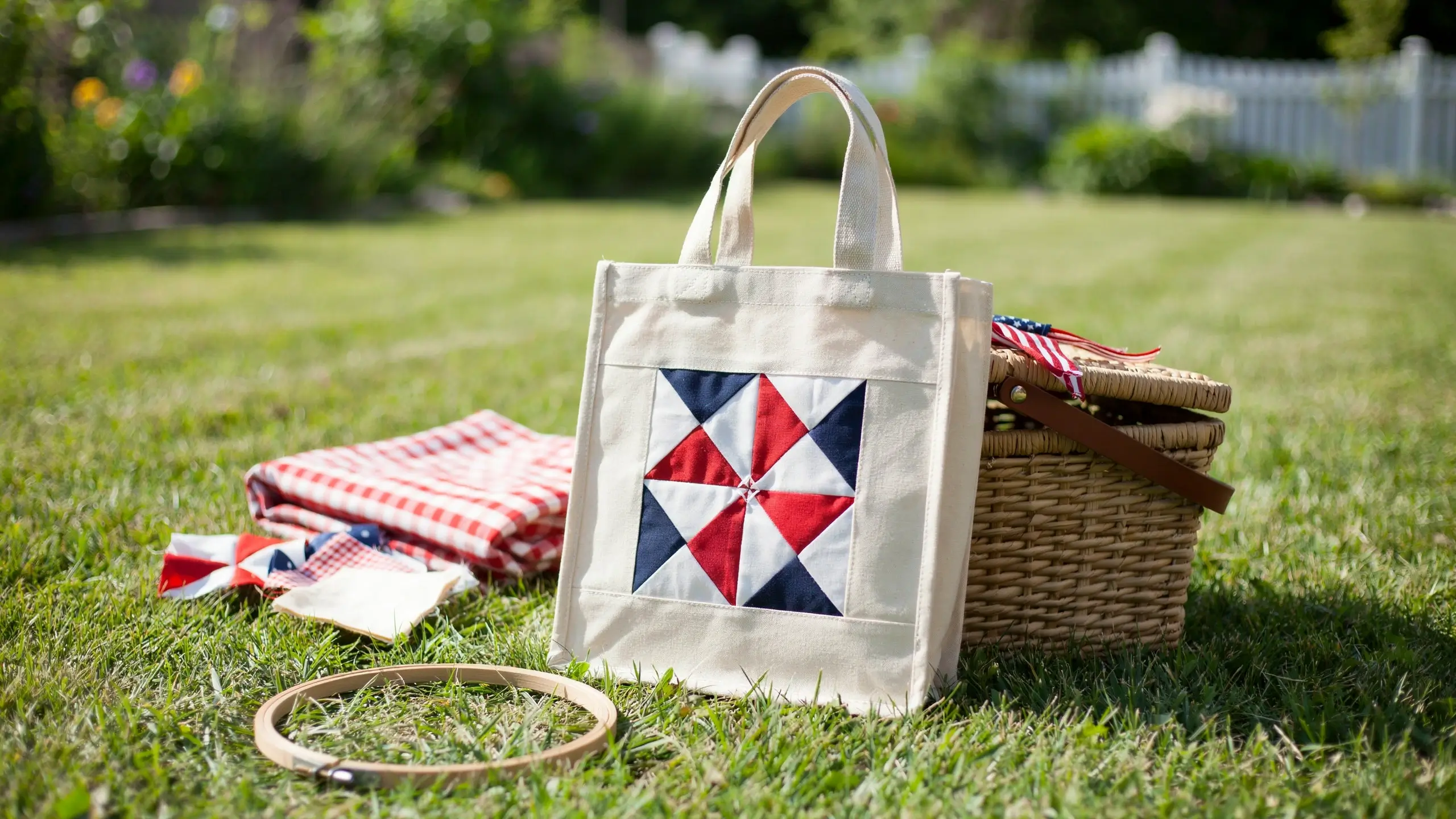 A cream canvas tote bag featuring a patriotic red, white, and blue pinwheel quilt block sits upright on a sunny grassy lawn beside a woven wicker picnic basket, folded red and white checkered cloth, and a wooden embroidery hoop, with a soft-focus white picket fence in the background