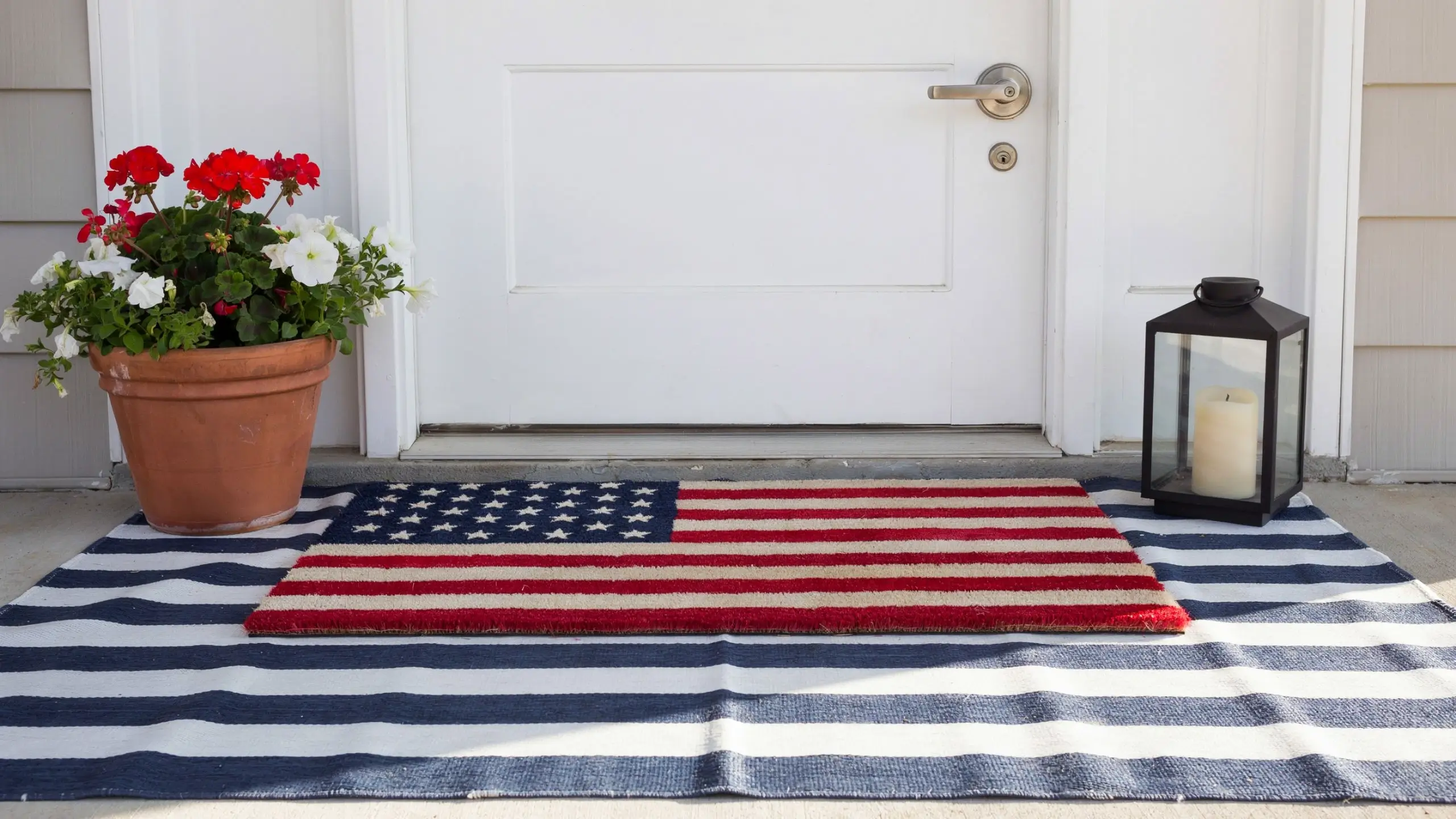 American flag doormat layered over a navy-and-white striped rug beside flowers and a lantern.