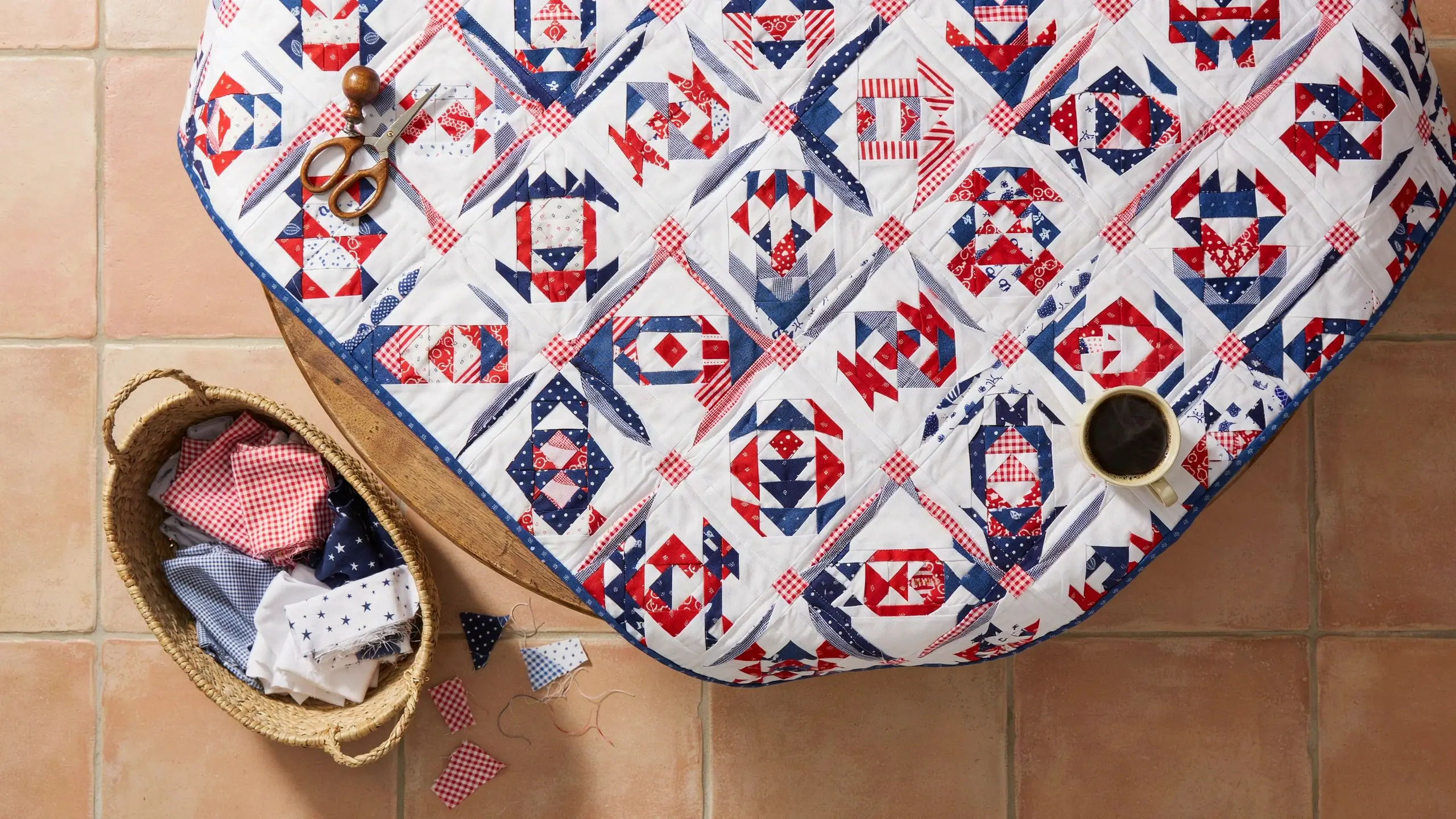 Handmade red, white, and blue Patriotic Pineapple quilt on a wooden table with a coffee mug and sewing scissors.