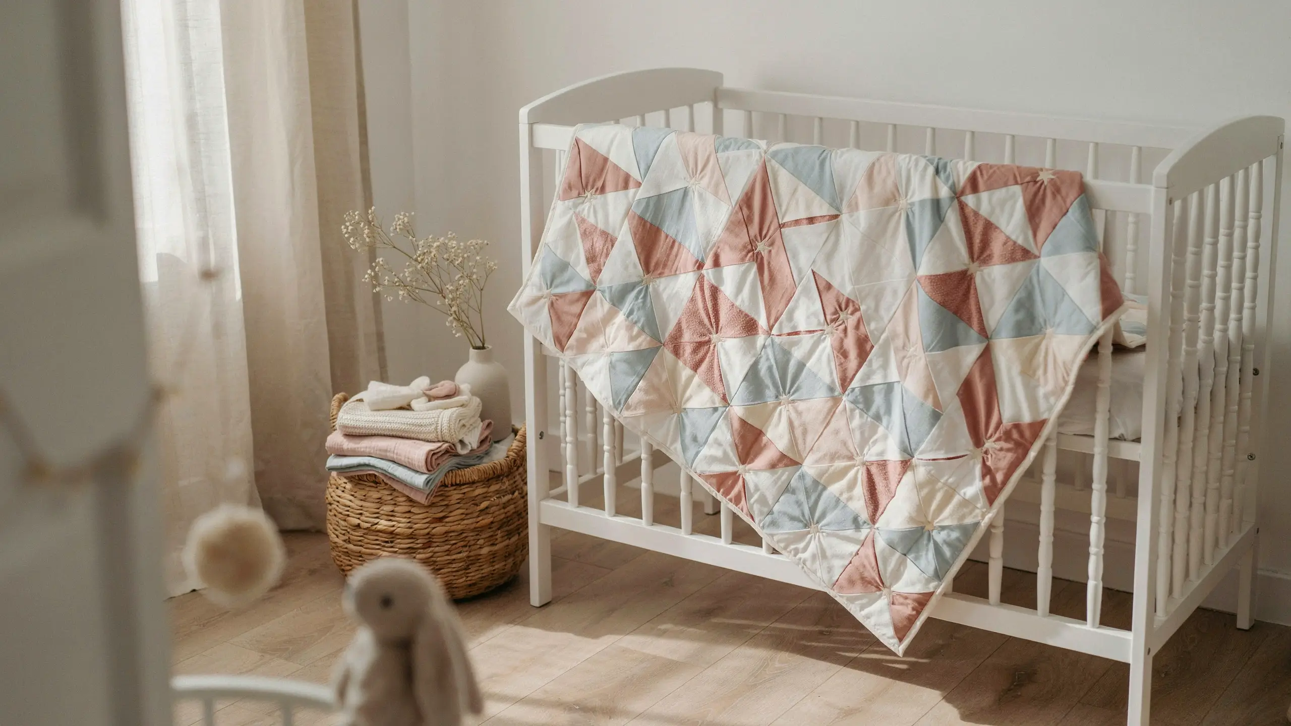 A soft patchwork baby quilt featuring a dusty rose, slate blue, and cream pinwheel star pattern, draped over a white wooden crib in a minimalist, sunlit nursery with a woven basket and a plush bunny toy.