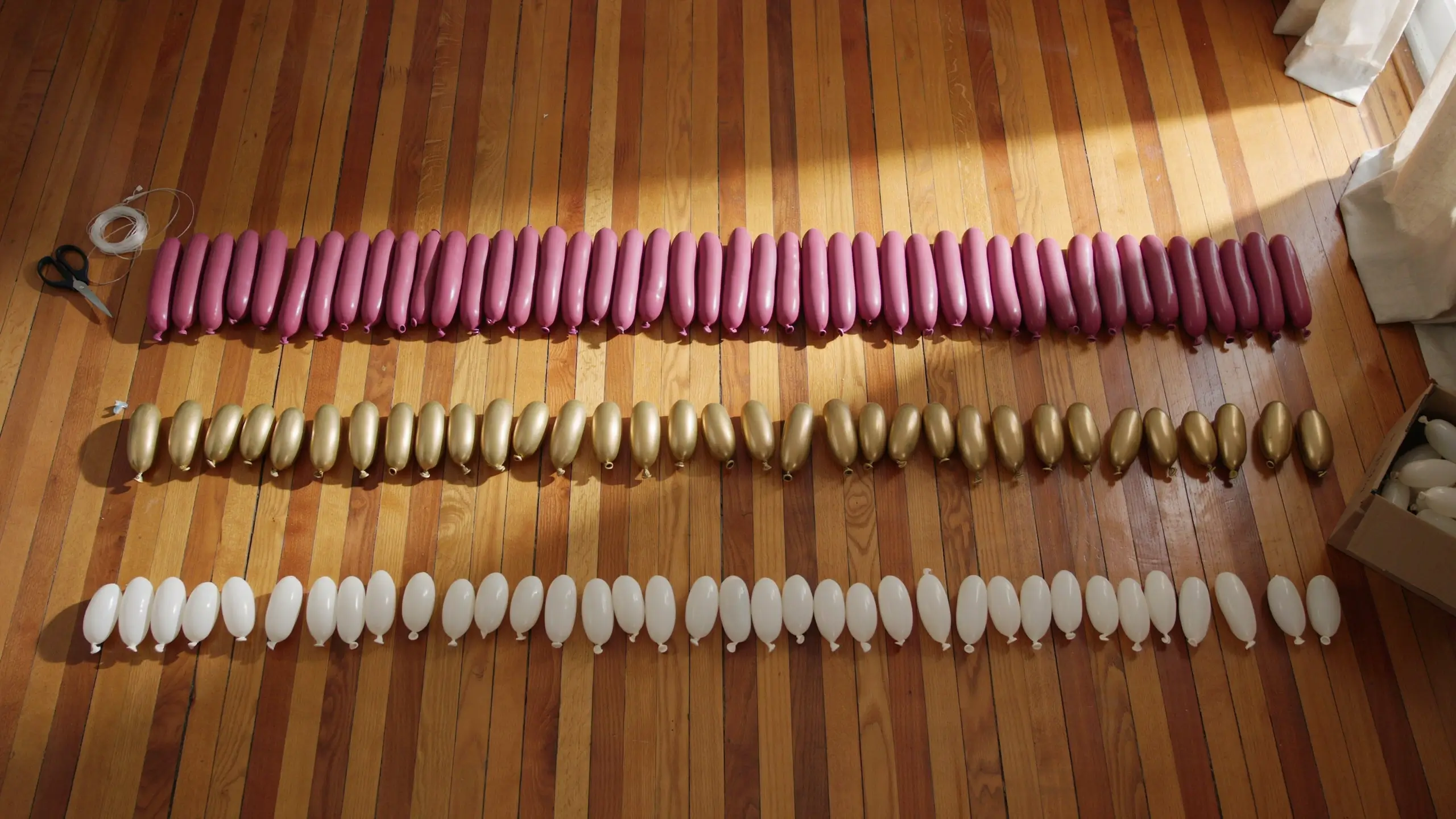 Flat lay of uninflated magenta, metallic gold, and white party balloons arranged in separate rows on a polished wooden floor with scissors and string, illustrating step one: planning the event color palette