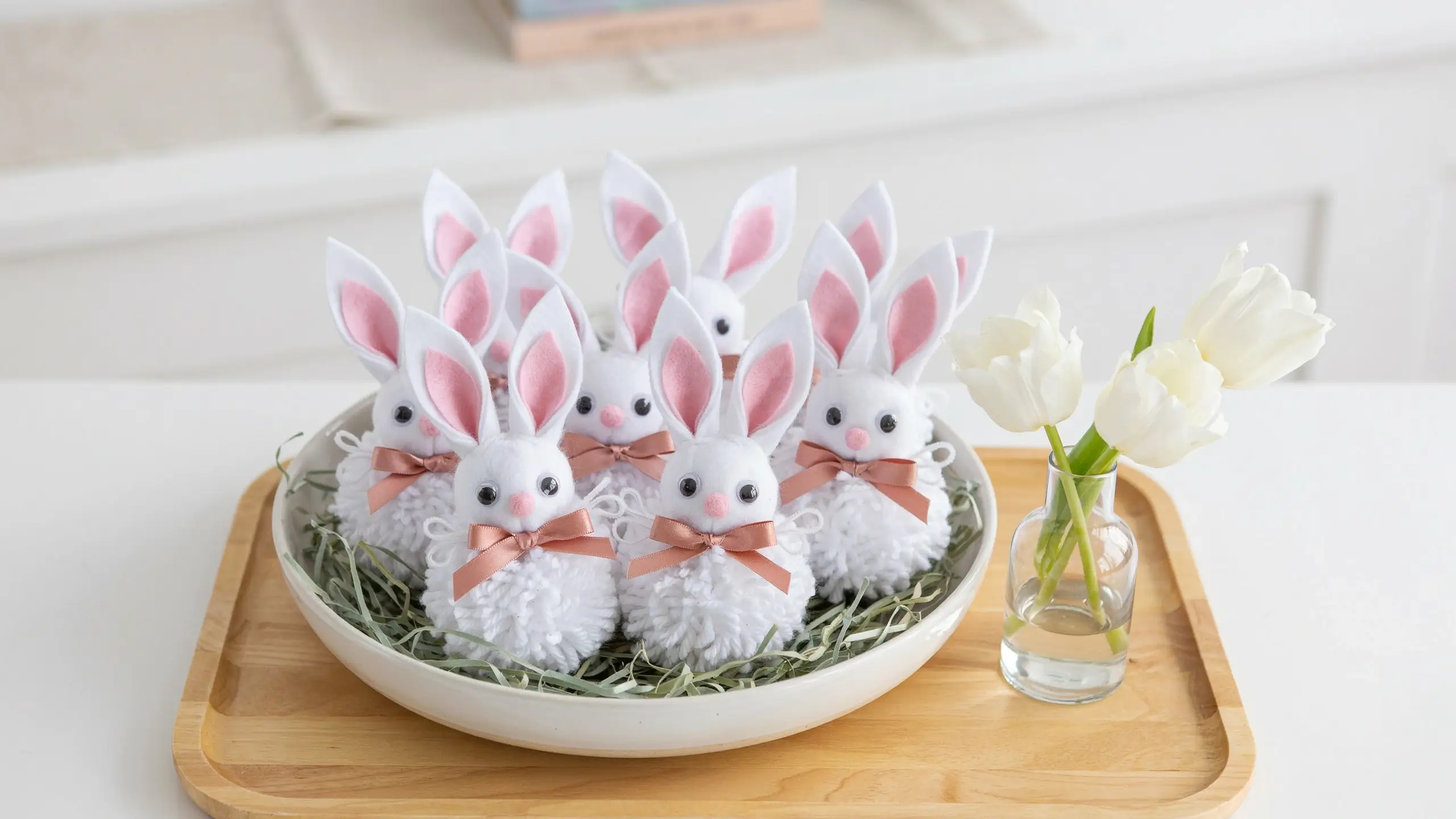Handmade white pom-pom Easter bunnies with pink felt ears and rose-gold bows sitting on a wooden tray with a glass vase of white tulips. 