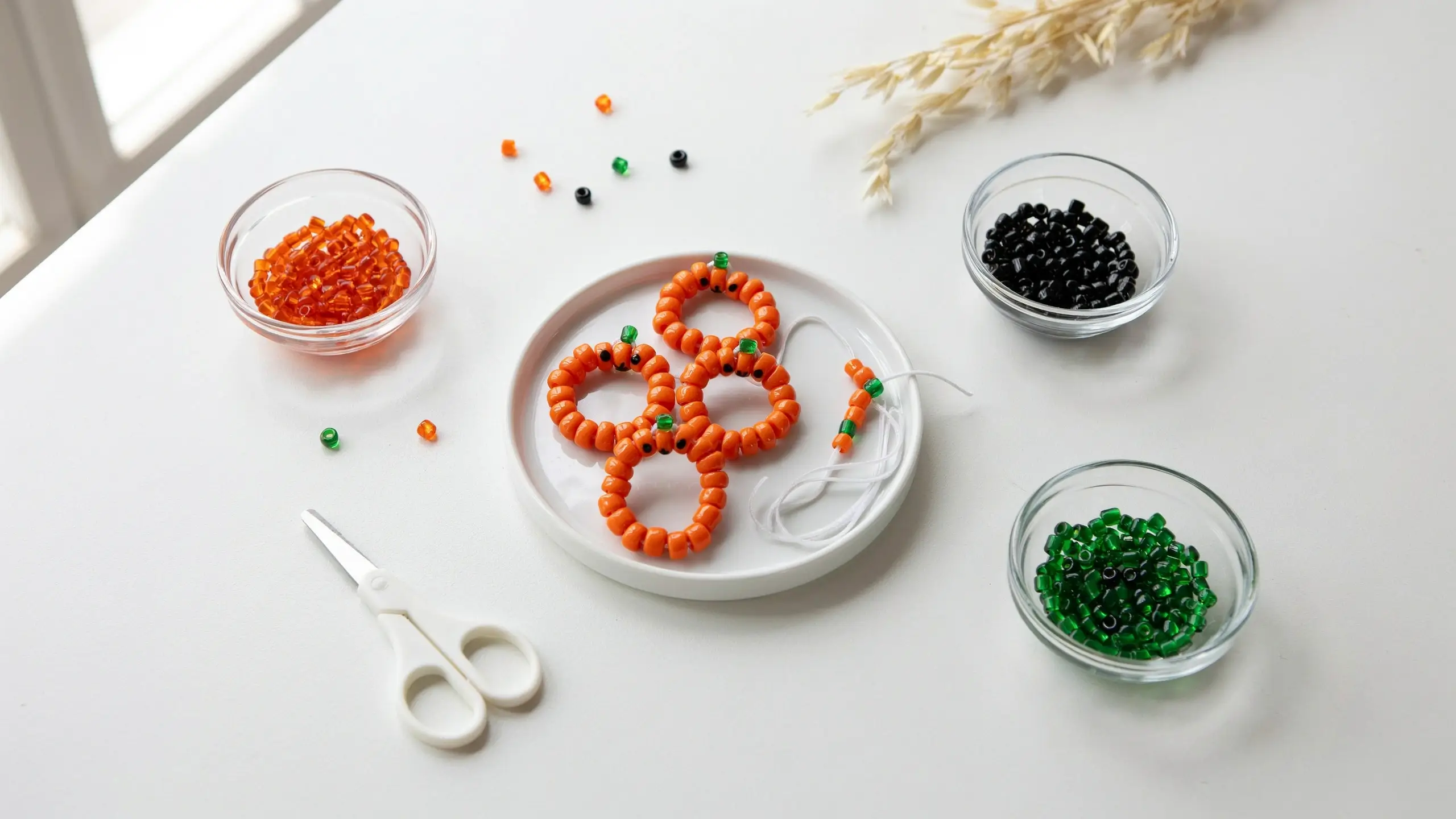 Close-up of three completed pony bead pumpkin bracelets featuring black eye and green stem beads resting on a white plate, surrounded by clear glass bowls of orange, green, and black pony beads, white crafting string, and scissors on a white table with bright natural daylight.